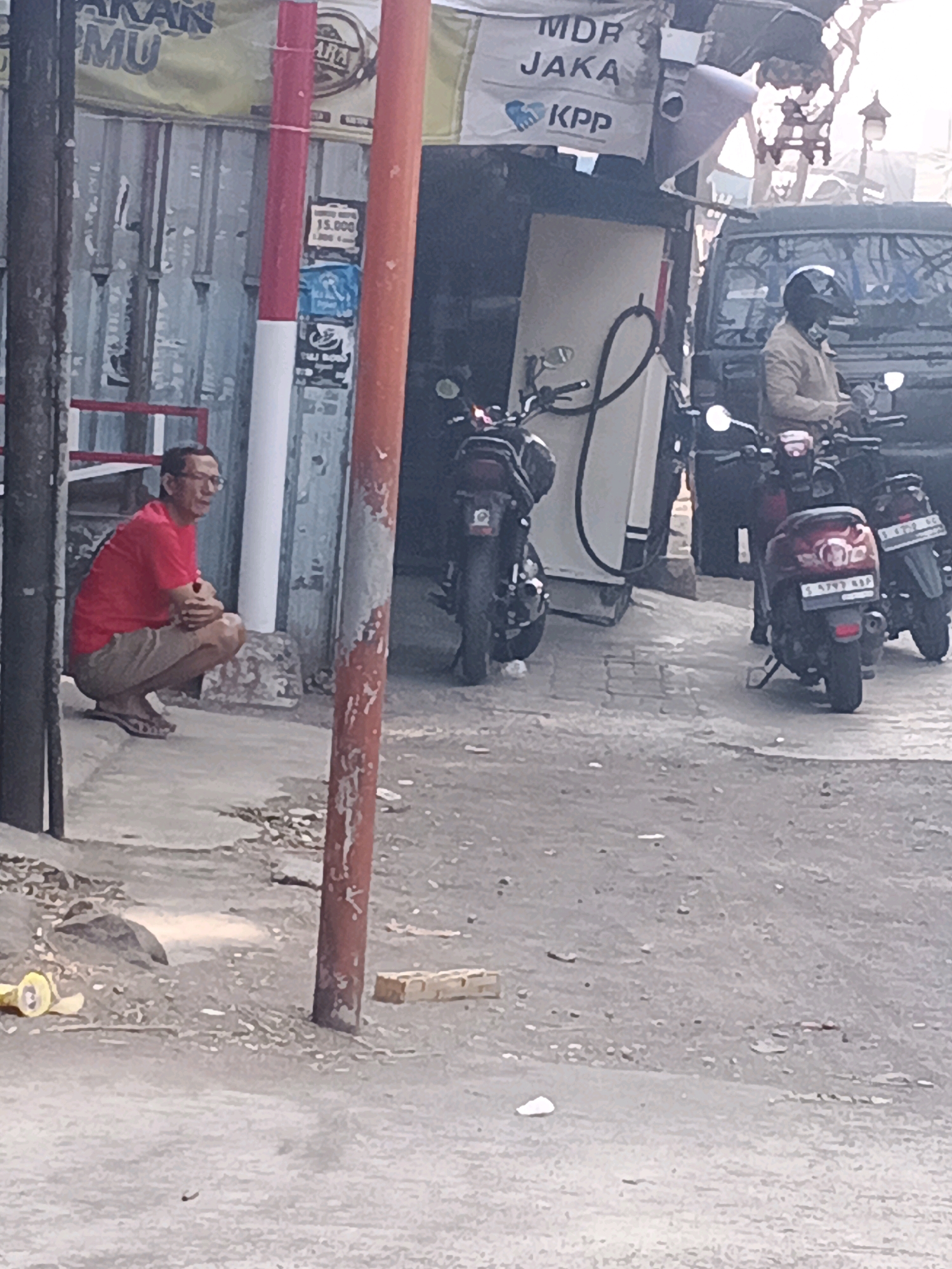 A man wearing glasses, a red t-shirt, and tan shorts squats on the sidewalk in Tambak Kemerakan, Indonesia. He is facing left, with his hands clasped in front of him. In the background, there is a metal wall, a red and white pole, and a gas pump. Several motorcycles are parked nearby, and a man wearing a helmet is sitting on one of them. A truck is also visible. The ground is paved with asphalt.