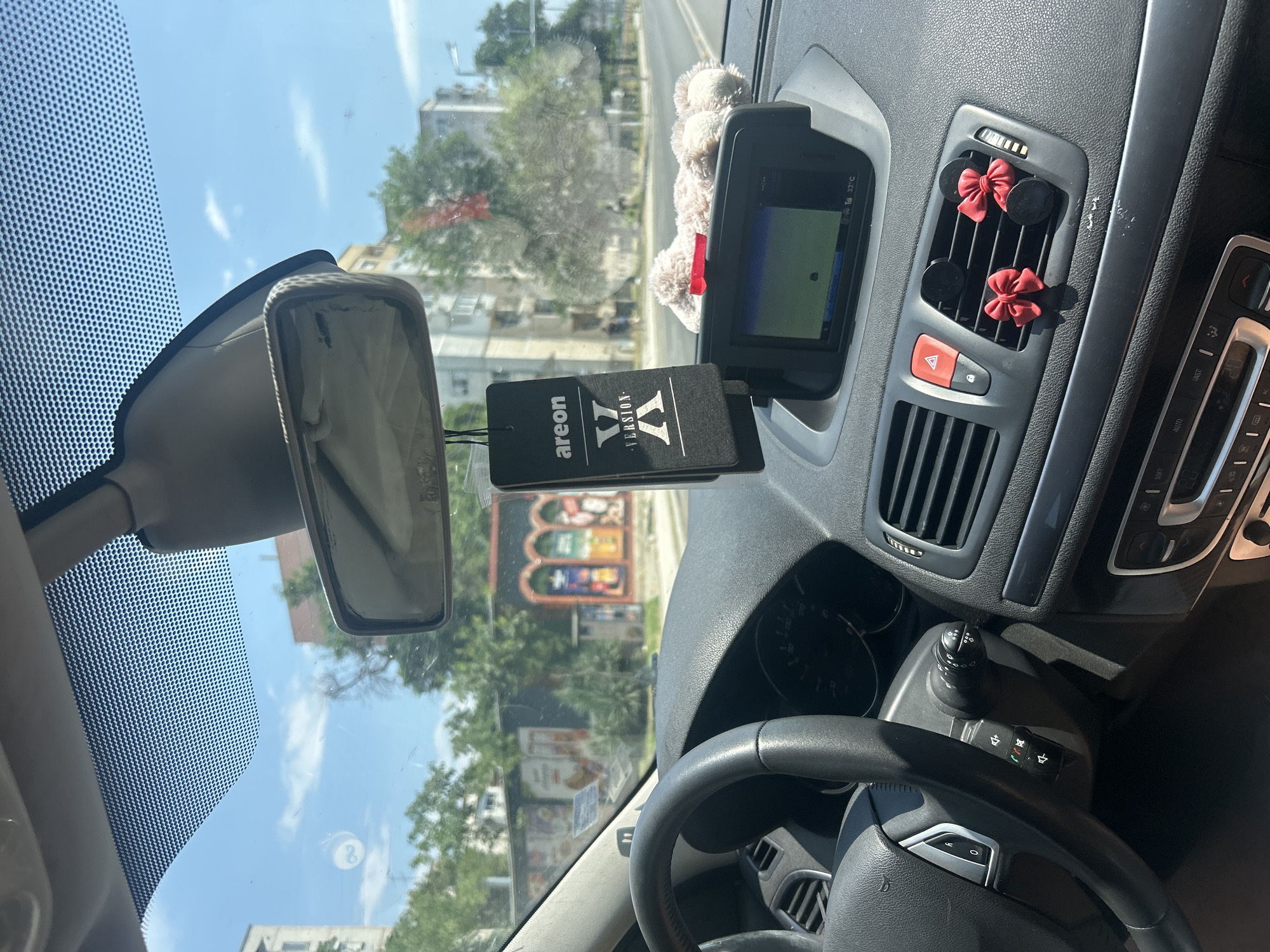 The image shows the interior of a car, likely parked on a street in Plovdiv, Bulgaria. The view is from the passenger seat, looking towards the dashboard and windshield. The dashboard features a steering wheel on the left, a central navigation screen, air vents, and various controls. A black "Areon X Version X" car air freshener hangs from the rearview mirror. A plush toy, possibly a teddy bear, rests on the dashboard near the navigation screen. Two small red bow decorations are attached to the air vents. Outside the car, buildings and trees are visible, suggesting an urban environment. The sky is blue with scattered clouds. The image appears to have been taken during daylight.