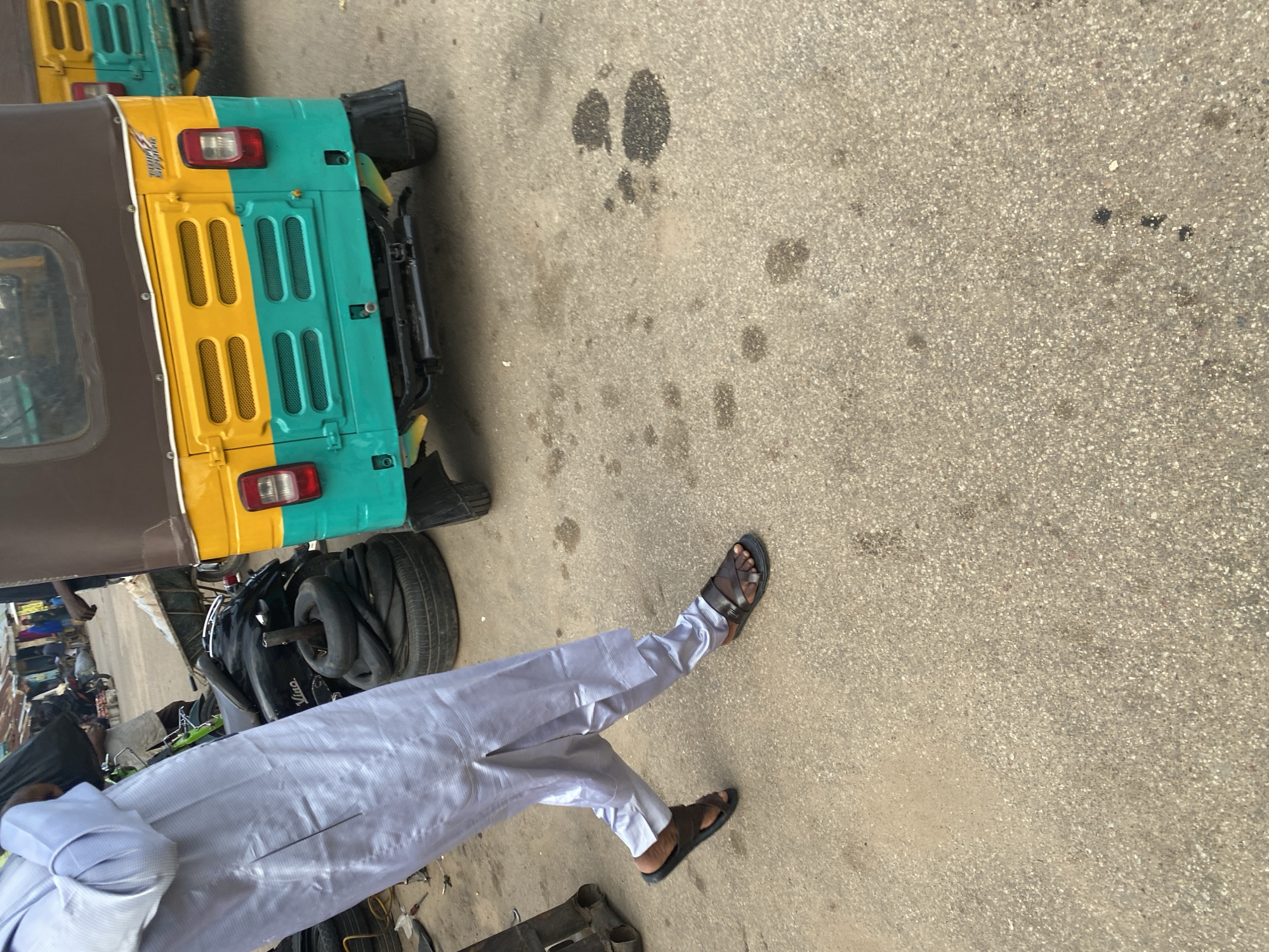 The image shows a close-up view of a person's legs and feet wearing white traditional attire and dark sandals, standing on an asphalt surface. The person is positioned on the right side of the frame, with their legs extended and slightly bent. In the background, the rear section of a yellow and turquoise auto-rickshaw is visible, along with some spare tires. The auto-rickshaw appears to be parked or stationary. The asphalt surface has some dark spots, possibly oil stains or other debris. The overall scene suggests an outdoor setting, likely a street or roadside in Jos, Nigeria, given the context. The lighting implies it is daytime, and there are no strong indicators of specific weather conditions. The image focuses on the person's lower body and the immediate surroundings, without revealing their face or a broader view of the environment. There is no visible text in the image.