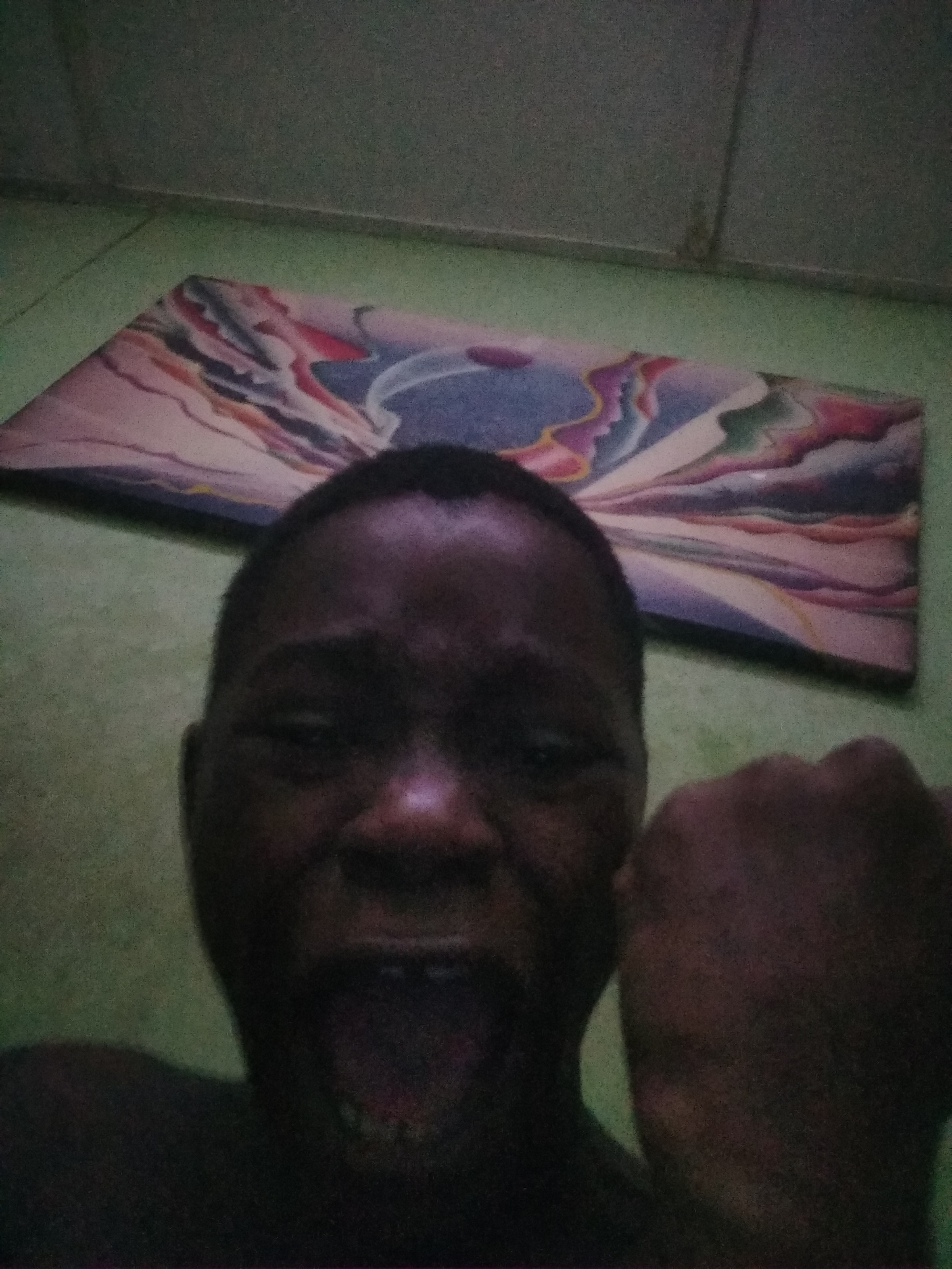 A close-up selfie of a dark-skinned individual with a shaved head and teeth showing, potentially sticking out their tongue, holding a fist up. A painting or poster with abstract swirling colorful designs lays on the floor behind them. The wall and floor appear to be of a light green or turquoise color. The location is not discernable as Shangisha, Nigeria from the image.