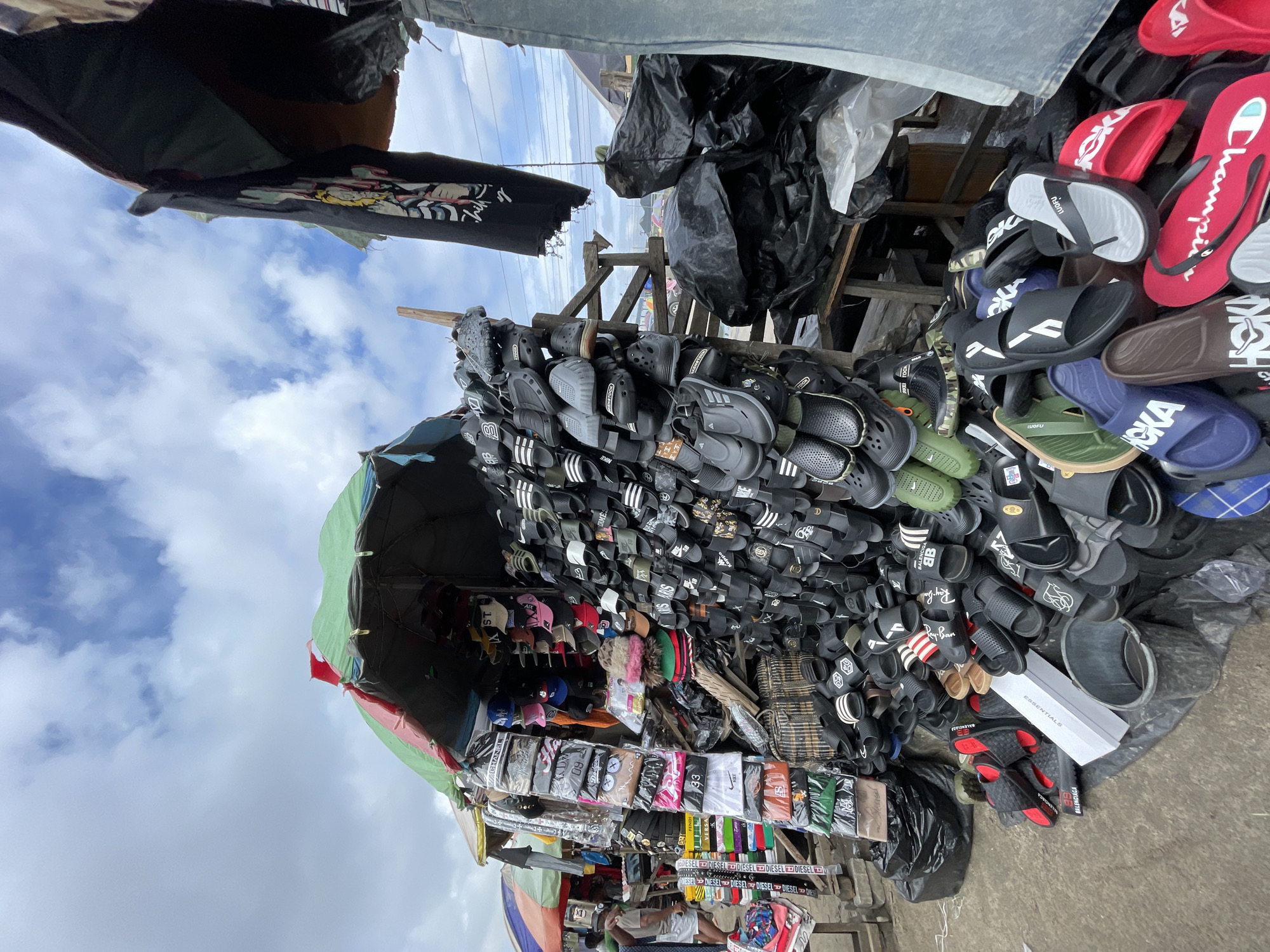 A street market scene in Lekki, Nigeria, features a vendor's stall overflowing with footwear. Black and white flip-flops, some with red stripes, are predominantly displayed on racks and piled on the ground. Various styles of sandals and slides, including Crocs-style footwear and slides with brand logos like "Balenciaga" and "Fendi," are visible. Some footwear is presented in their original packaging. Among the footwear, stacks of hats, including baseball caps, are arranged on shelves. A black t-shirt with a graphic print hangs from a wooden beam above the stall. Behind the stall, large black trash bags are piled, and some tents and structures are visible in the background. The ground is paved, and the sky above is partly cloudy. A person in the background is partially visible, appearing to be a customer.