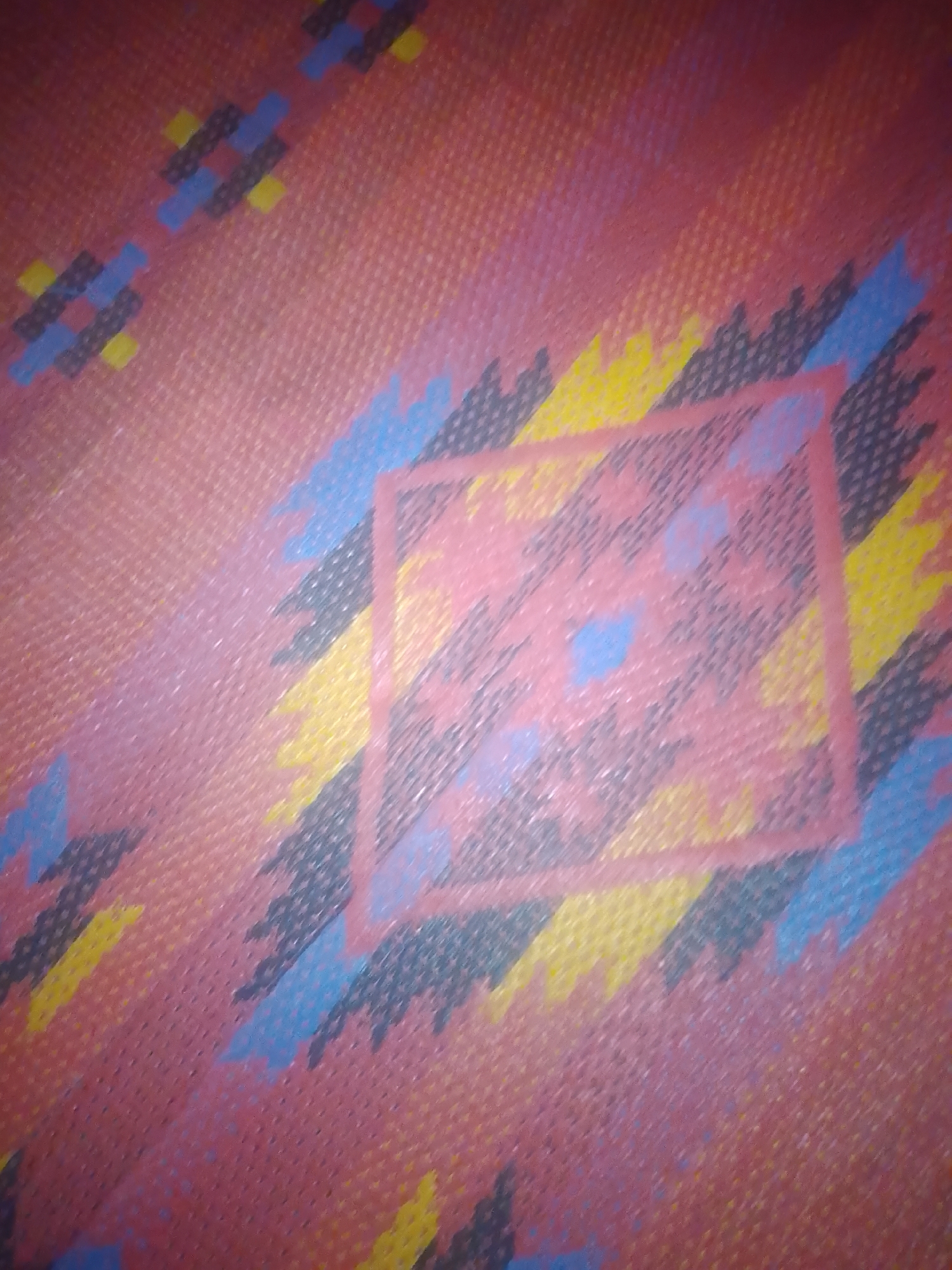 The image displays a close-up view of a patterned textile, likely upholstery or fabric. The dominant color is a reddish-pink, textured with a fine weave visible throughout. Overlaying this background are geometric shapes and designs in various colors, including black, yellow, and blue. These patterns appear abstract and fragmented, with diagonal lines and jagged elements creating a dynamic visual effect.
There is a prominent square or rectangular frame, outlined in a lighter pinkish hue, centrally positioned within the image. Inside this frame, the pattern is more concentrated and features smaller blue shapes resembling dots or irregular splotches. The overall impression is of a decorative textile with a somewhat retro or ethnic-inspired design.
There are no people or identifiable landmarks visible in the image. The photograph is taken from a fixed perspective, focusing solely on the surface of the fabric. There is no indication of the specific location in Jauro Yinu, Nigeria, or any context that would suggest its placement within the city.
