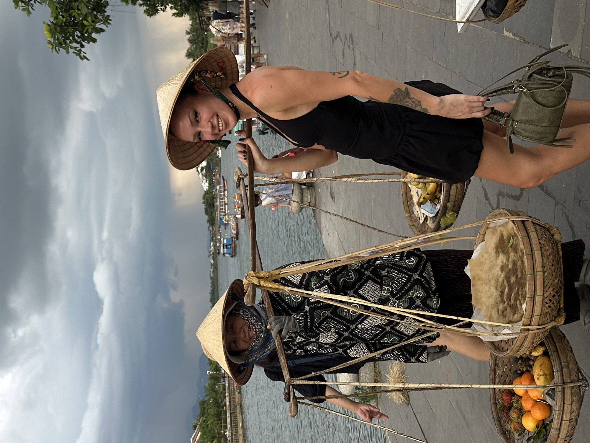 The image shows two women, one of Vietnamese ethnicity and the other appearing to be a tourist, standing on a paved walkway next to a body of water in Hoi An, Vietnam. The Vietnamese woman, on the left, wears a conical hat and a patterned shirt, carrying a shoulder yoke laden with baskets of fruit, including oranges, rambutans, and what appear to be mangoes and bananas. The tourist, on the right, also wears a conical hat and a black dress, holding a green handbag and a shoulder yoke with baskets. Behind them, boats are visible on the river, and other people are walking along the waterfront. The sky is overcast with dark, dramatic clouds, suggesting a storm is approaching. The scene depicts a common sight in Hoi An, where traditional vendors sell goods along the riverbanks.