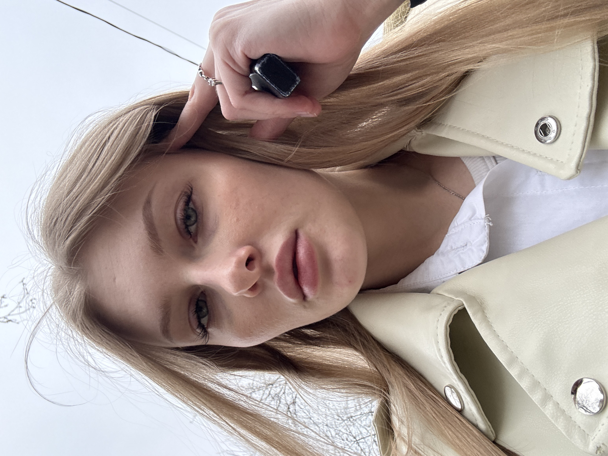 A portrait of a blonde woman with fair skin, wearing a white top and a light yellow jacket. She is positioned with her face close to the camera, tilted slightly. Her left hand is visible near her head, holding a small, dark object. She is wearing a ring on her finger. The background shows a cloudy sky and indistinct outlines of trees, possibly indicating an outdoor setting. No specific location information is discernible from the image.