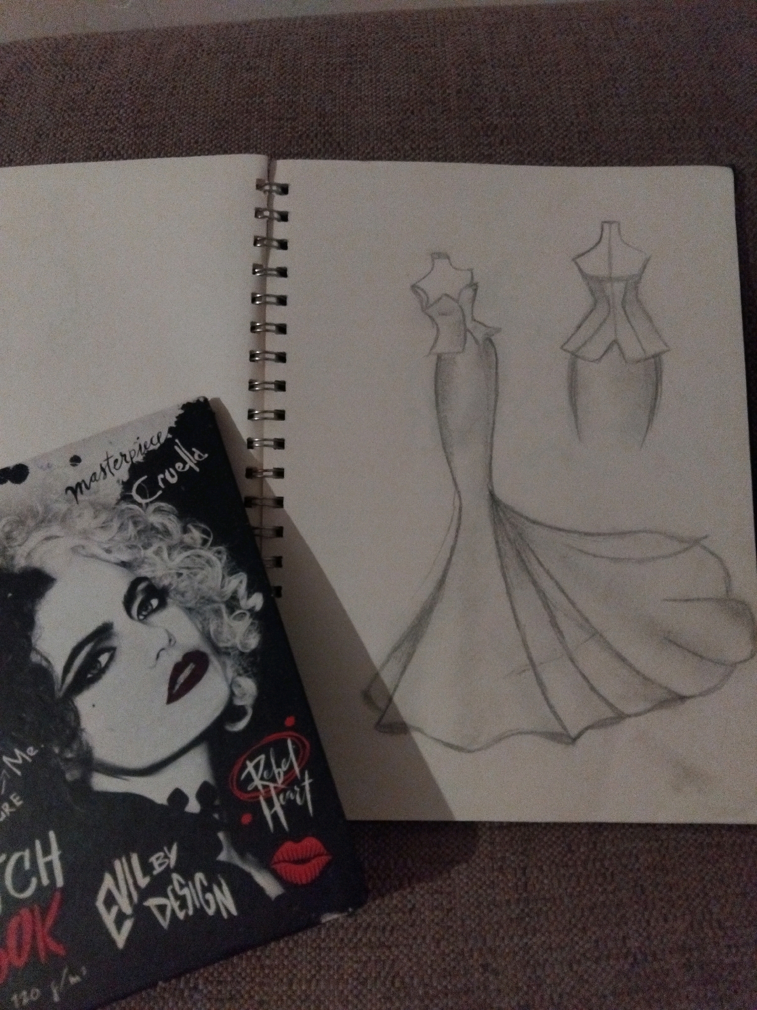 A sketchbook lies open on a textured brown surface, likely a couch or carpet. On the left page, a portion of a book cover is visible, featuring a black and white illustration of a woman's face with dramatic makeup, curly hair, and red lips. Text on the book cover includes "masterpiece Cruella" and "EVIL BY DESIGN". The right page of the sketchbook contains two pencil sketches of dresses. The larger sketch on the right depicts a full-length gown with a fitted bodice and a dramatic, flowing train. A smaller sketch to the upper right shows the bodice of a different dress, with a distinct peplum detail. The scene appears to be indoors, with natural light illuminating the sketchbook.