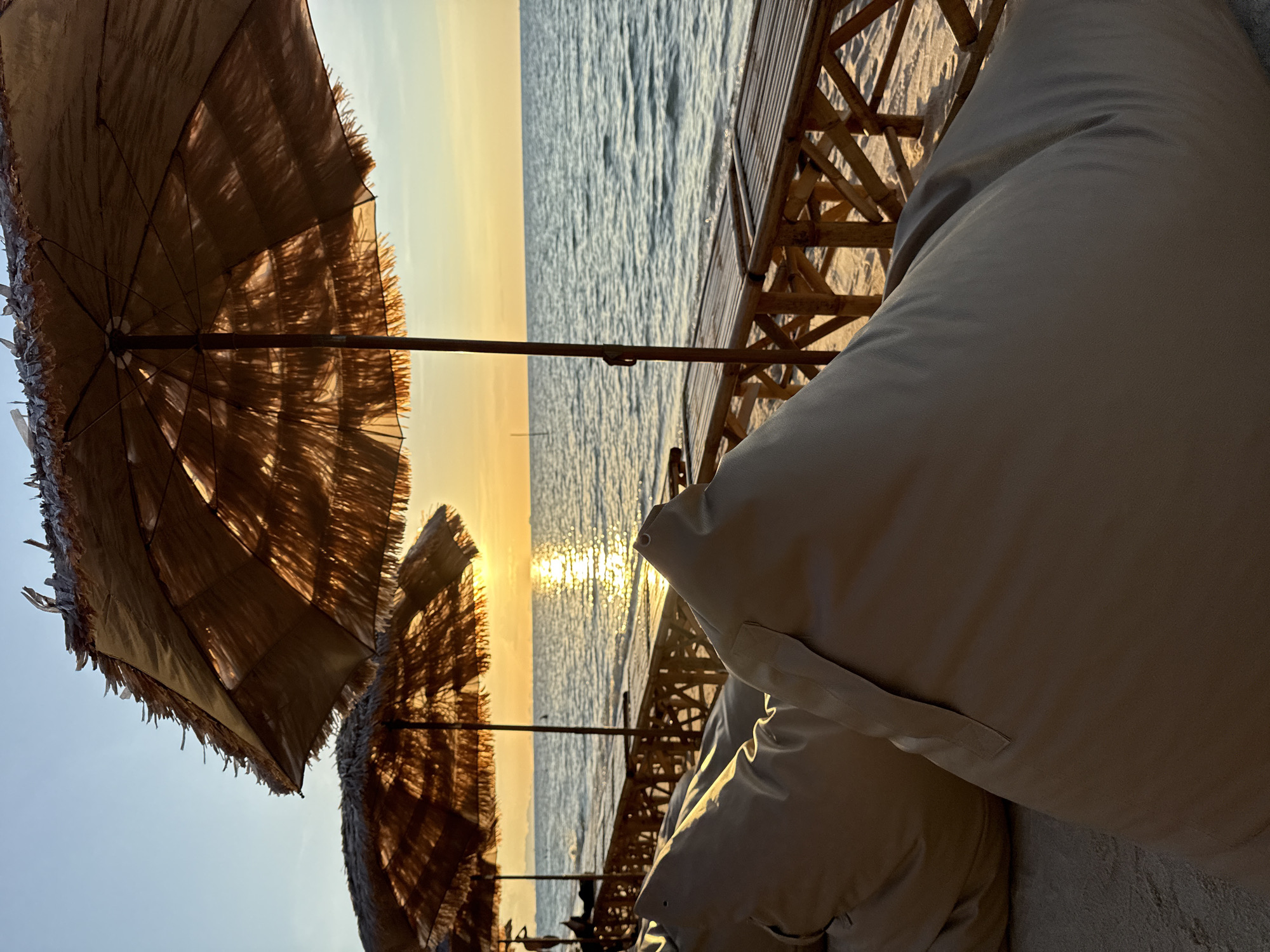 The image depicts a beach scene in Baan Tai, Thailand during sunset. In the foreground, two large, light-colored beanbag chairs rest on the sand. Behind them, a row of bamboo-slatted lounge chairs is visible, some occupied by figures. Several thatched umbrellas provide shade. The calm sea stretches to the horizon, reflecting the warm hues of the setting sun. The sky is a gradient of pale blue near the top, transitioning to soft orange and yellow near the horizon.