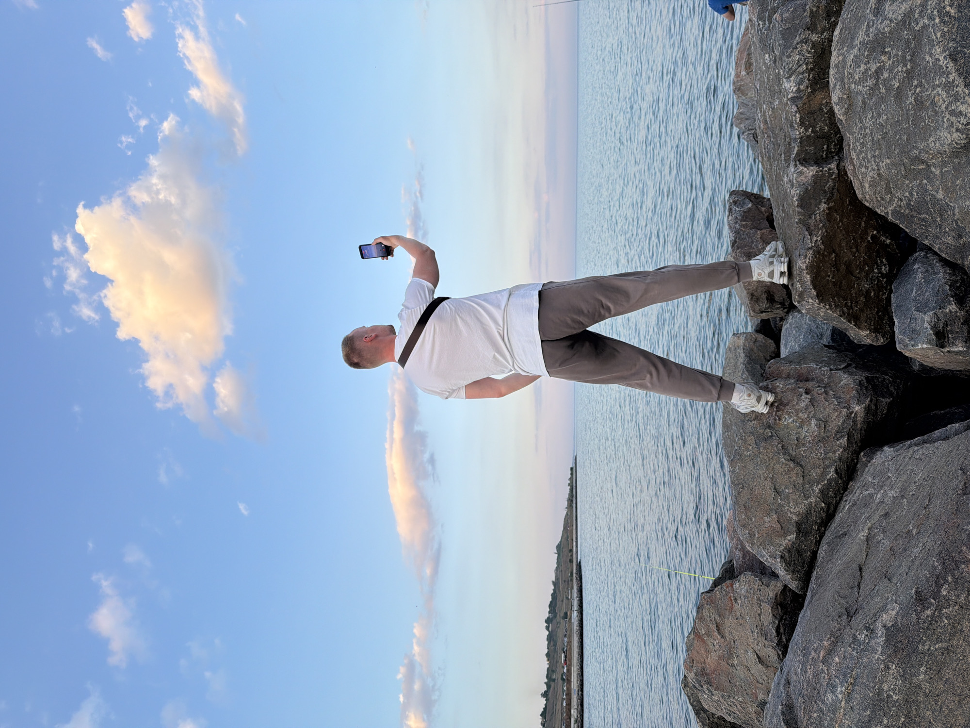 A man stands on large, dark grey rocks on the coast near Fontanka, Ukraine, facing away from the camera and towards the sea. He is wearing a white t-shirt, grey sweatpants, and white athletic shoes. A dark strap is visible across his t-shirt. In his right hand, he holds up a black smartphone, appearing to take a selfie or record a video. The sea is calm and blue, stretching to the horizon. The sky is a pale blue with scattered white and pink-tinged clouds. In the distance, a long, thin line of land with some structures and parked cars is visible along the coast. A fishing rod with a yellow line is planted in the rocks in the foreground.