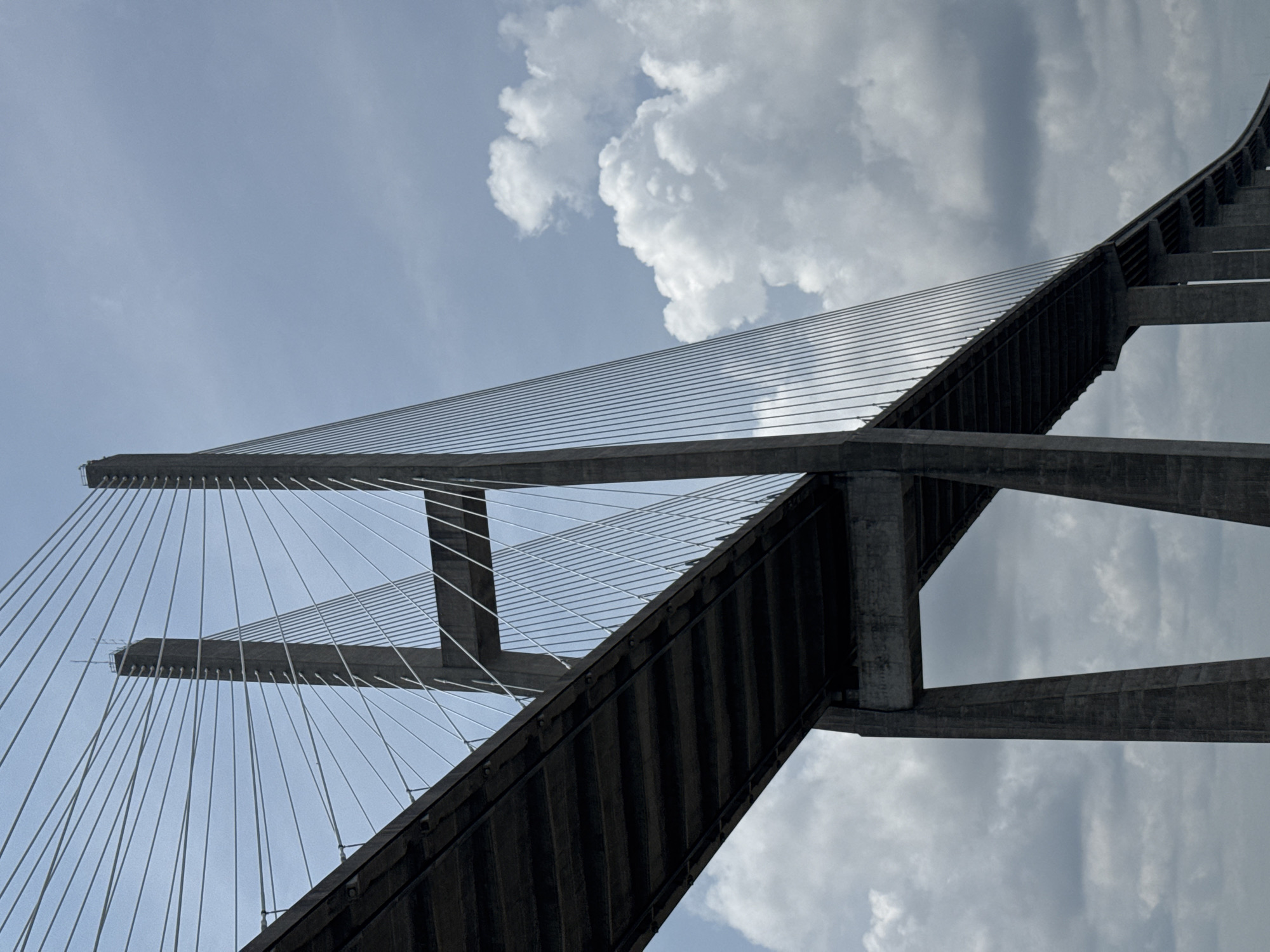 The image shows a low-angle perspective of the Sidney Lanier Bridge in Brunswick, Georgia, USA. The bridge is a cable-stayed bridge with two tall concrete pylons. The road deck of the bridge is visible in the foreground, extending from the left side of the image towards the right. Numerous white cables run from the tops of the pylons to the road deck, creating a striking geometric pattern against the cloudy sky. The pylons are massive concrete structures with a stepped design. The sky is partly cloudy, with a mix of bright white cumulus clouds and darker, more diffused clouds. The overall impression is one of immense scale and engineering prowess.