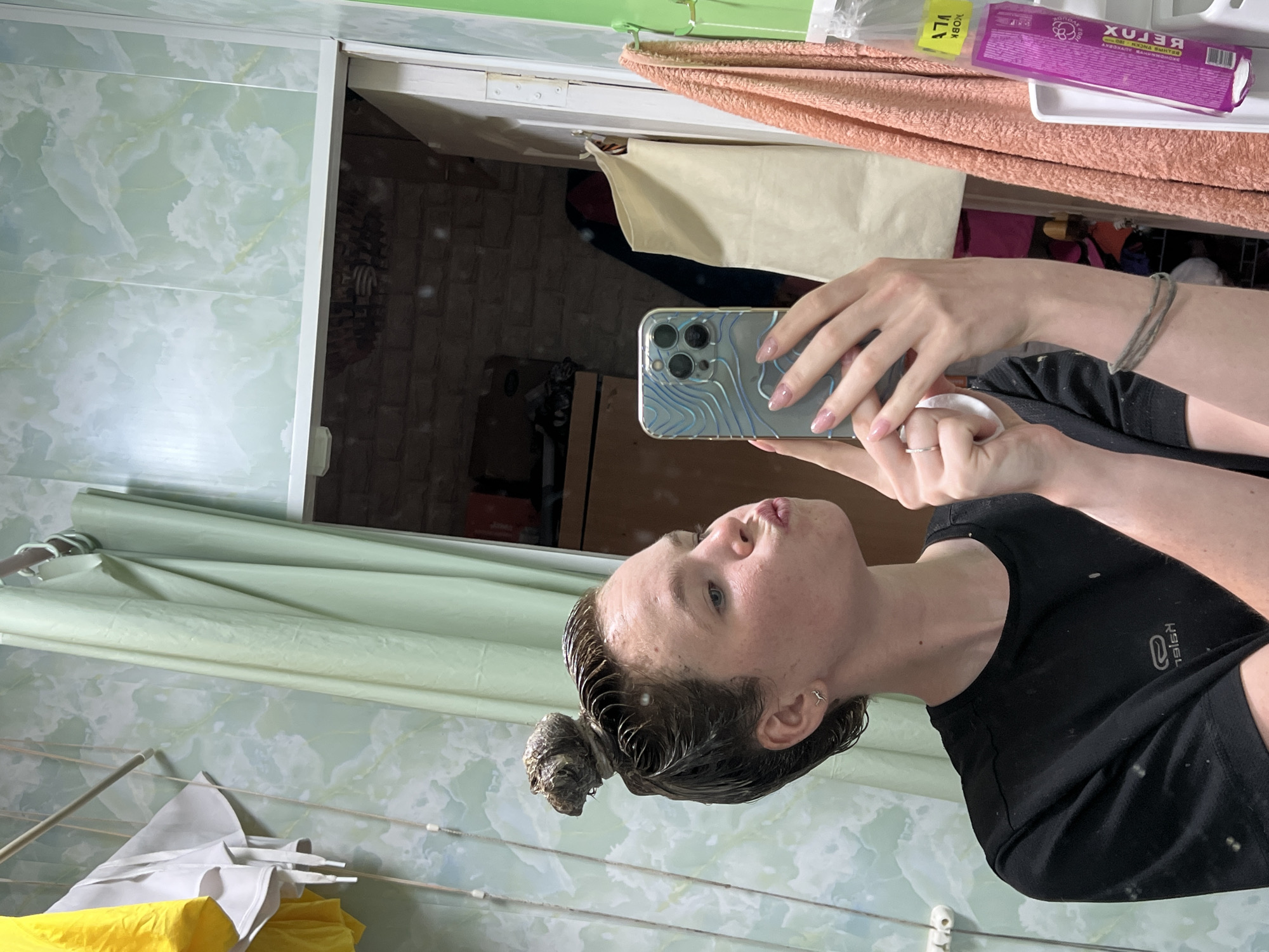 A woman is taking a selfie in a bathroom. The woman is wearing a black shirt and has dark, wet hair pulled up into a bun on top of her head. She is holding a smartphone in her left hand, capturing her reflection in a mirror.
The background includes the bathroom environment. The walls are covered in light green marble-patterned tiles. A white cabinet is visible above, and a peach-colored towel and a purple box of wipes rest on top of it. The reflection in the mirror shows a storage unit or shelves with miscellaneous objects inside. Below the woman, a clothing rack holds white and yellow garments. The scene appears to be in a residence.
Based on the presence of clothing rack, the individual may live in an apartment or house, not a hotel or other transient accommodation.
While it is possible to infer the location based on the Russian text on the wipes, this image cannot verify that the photograph was taken in Chaikovsky, Russia.