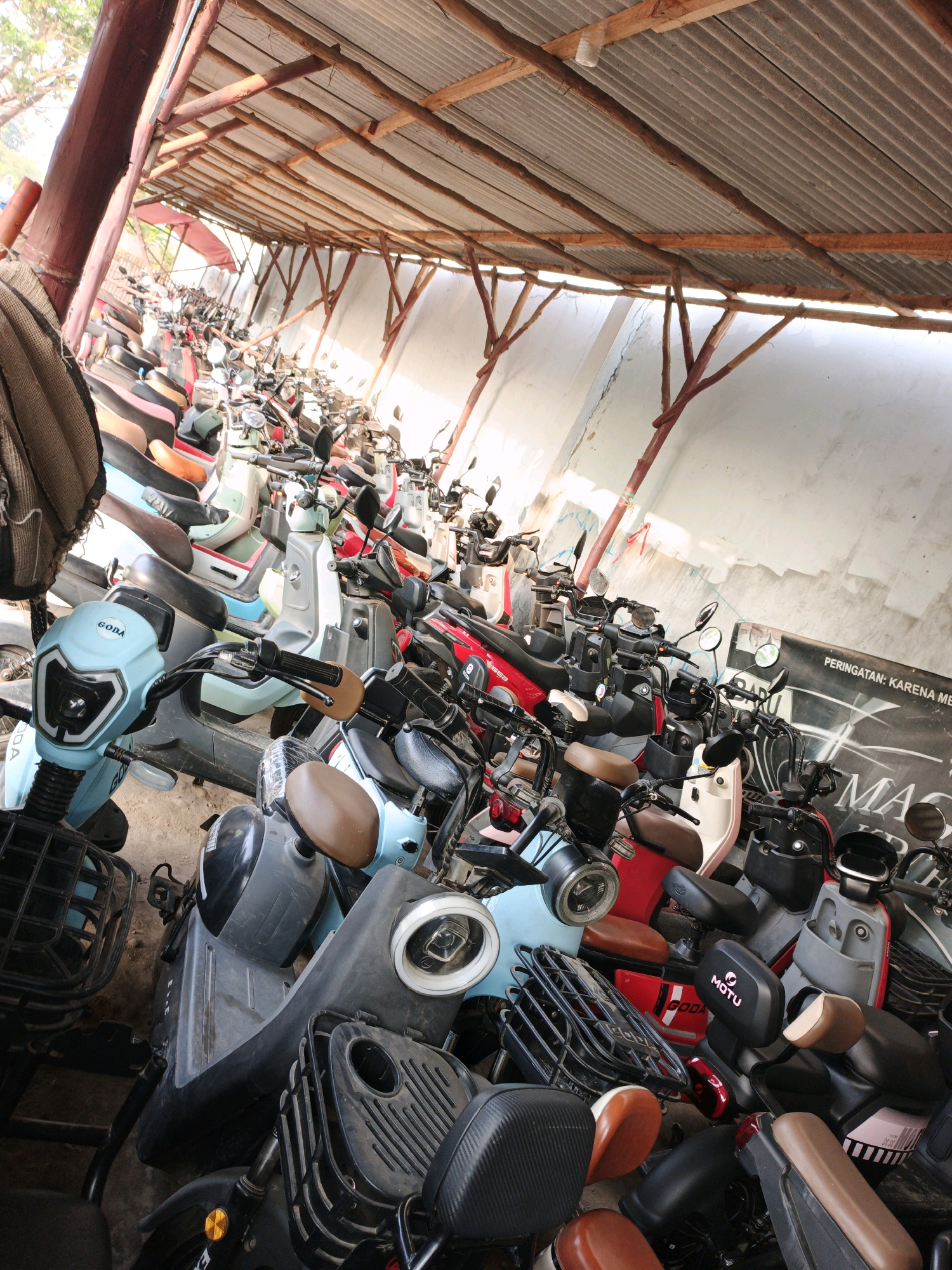 The image shows a large outdoor parking area, possibly a storage lot or charging station, filled with many electric scooters. The scooters are packed closely together under a corrugated metal roof supported by wooden beams. The lighting suggests it is daytime, with sunlight filtering through the gaps in the roof and from the open sides. A white wall, possibly a building exterior, forms the background.
The scooters are varied in color, including light blue, red, black, white, and grey. They appear to be new or well-maintained. Some have baskets or storage compartments in the front. Several scooters have visible branding, such as "GODA" and "MOTU."
There are no people visible in the image. The scene is static, depicting parked vehicles. The environment is utilitarian, designed for vehicle storage.
The location is identified as Krimun, Indonesia. This suggests the scene is a common sight in urban or semi-urban areas of Indonesia where electric scooters are utilized for transportation. The overall impression is one of a commercial or municipal facility dedicated to electric scooter storage or rental.