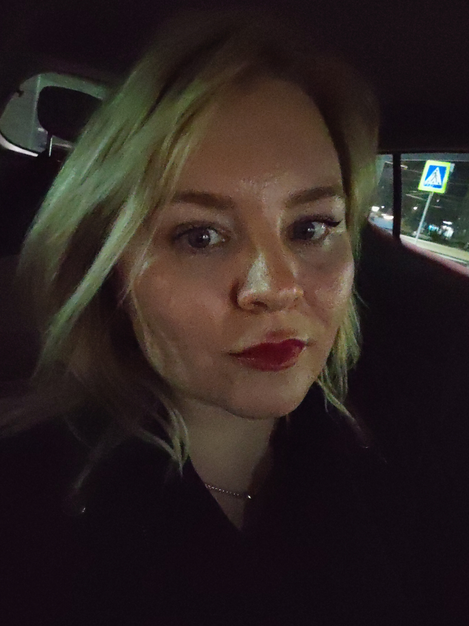 A close-up portrait shows a woman with blonde hair looking directly at the camera, captured at night inside a vehicle in Izhevsk, Russia. She has a neutral to slightly pouting expression, wearing dark clothing and a thin silver chain necklace. Her lips are adorned with red lipstick, and she has dark eyeliner.
The setting places her in the front or back seat of a car, with parts of the vehicle's interior, like a headrest and window frame, visible. Through the car window in the background, out-of-focus city lights and blurry shapes of buildings or trees suggest an urban environment at night. A distinct blue square road sign with a white triangle containing a white stylized "A" symbol is visible in the distance, characteristic of a pedestrian crossing or informational sign in Russia. The image has a noticeable grainy texture, indicating it was taken in low-light conditions.