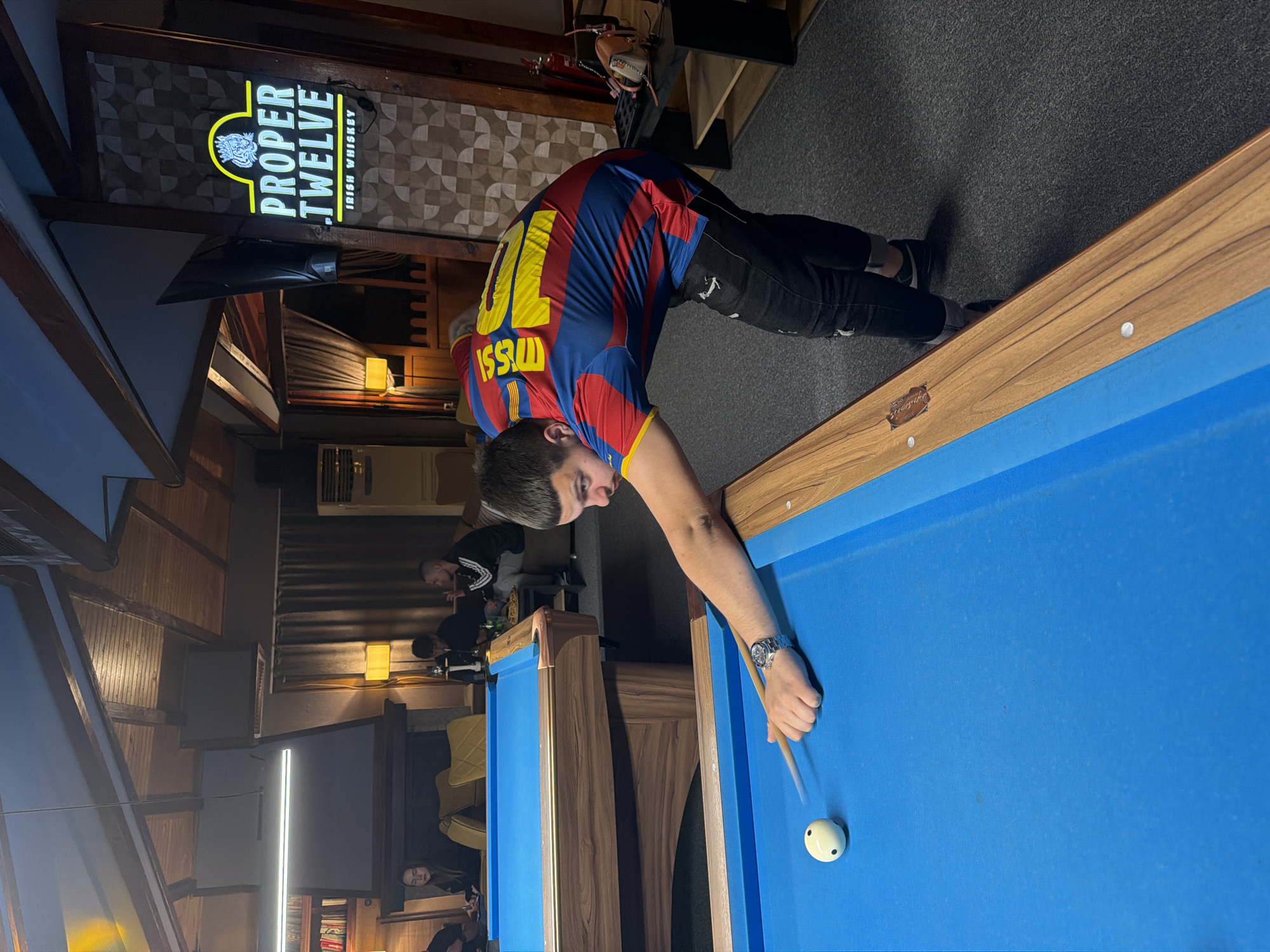 A man in a Messi #10 Barcelona jersey is playing pool on a blue pool table in a dimly lit establishment, presumably a bar or game room. He is bent over, cue stick in hand, aiming at a white cue ball. He wears a watch on his left wrist and black pants. In the background, other people can be seen sitting at tables. Above and to the left of the man is a "Proper Twelve Irish Whiskey" sign. The establishment is located in Plovdiv, Bulgaria.