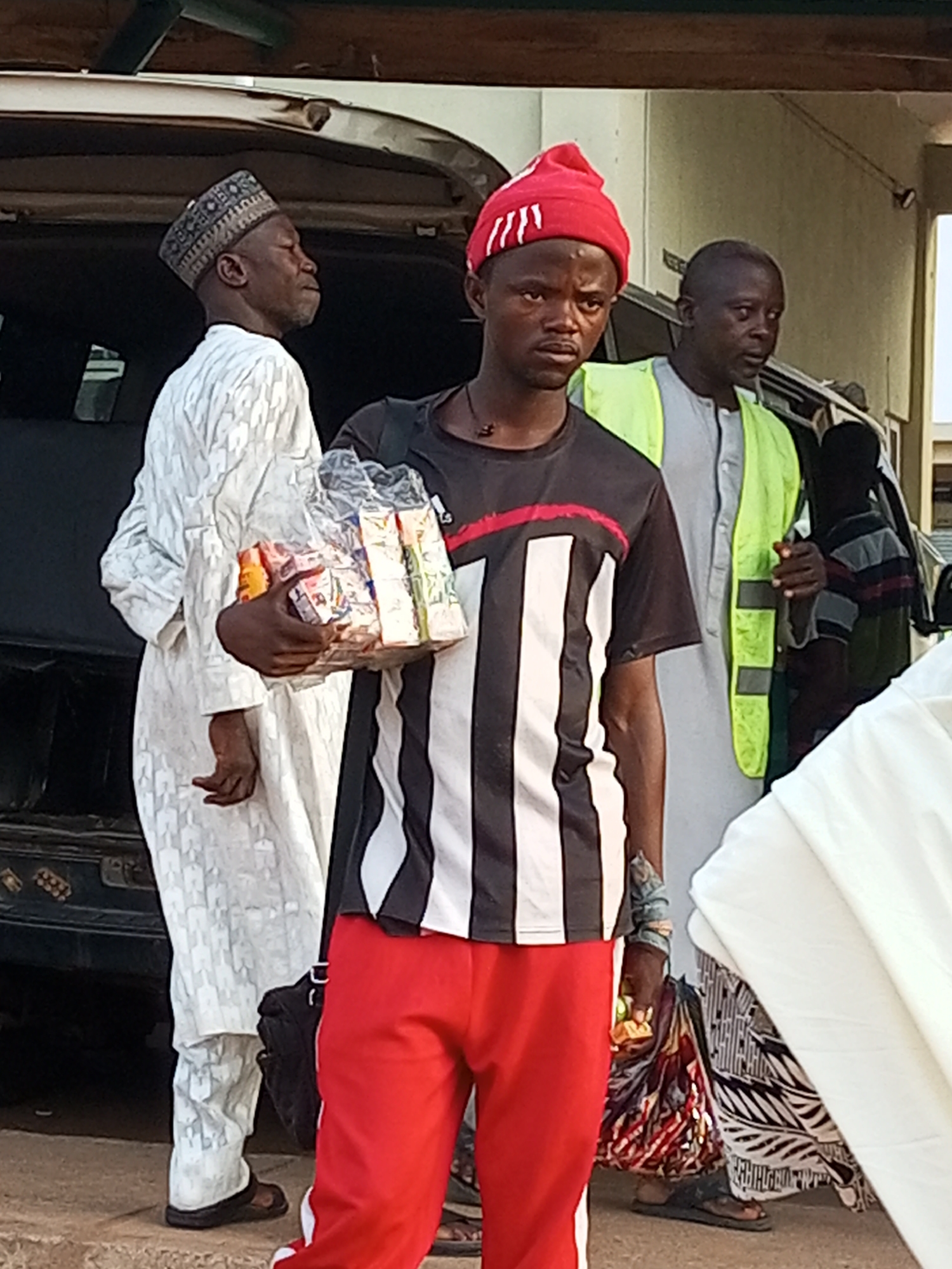 In Gombe, Nigeria, three men are gathered near the back of a dark-colored vehicle, possibly loading or unloading it. The man in the foreground wears a red beanie, a black t-shirt with white and red stripes down the sleeves, and red pants. He holds a clear plastic bag filled with food items, appearing to be snacks or packaged goods. To his left, a man in traditional white attire and a patterned cap looks towards the right. Behind the man in the red beanie, another man wears a light green high-visibility vest over a grey garment, looking to his right with a concerned expression. Further to the right, a fourth individual in a striped shirt is partially visible. The scene suggests an outdoor setting, possibly during the day given the lighting. The overall mood appears neutral, with the focus on the individuals and the items they are handling. No visible text is discernable in the image.