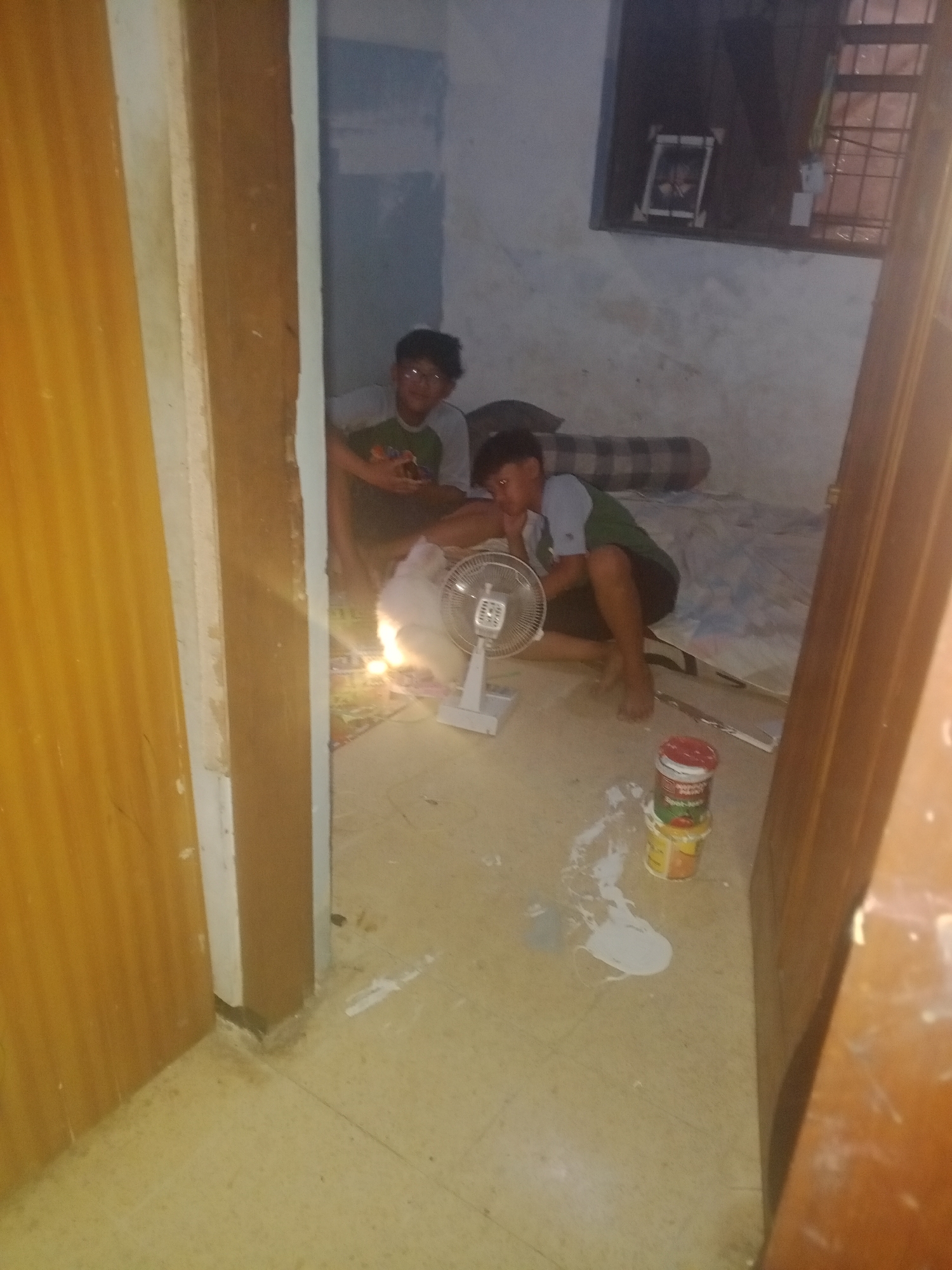 Two young males are present in a dimly lit, sparsely furnished room, potentially in Lembah, Indonesia. One male is seated on the floor, facing left, with his elbows on his knees and his head resting on his hand. He wears a grey and green t-shirt and dark shorts. In front of him, a white electric fan is operating, with a visible light source illuminating the area. The second male is partially visible behind him, also seated, and appears to be wearing similar clothing. The room has light-colored walls and a tiled floor. Several cans of paint, one with a red lid, are scattered on the floor, along with white paint splatters, suggesting a recent or ongoing painting activity. A striped bolster pillow lies on a bed or mattress in the background. A barred window is visible on the far wall. The overall impression is one of a makeshift living or work area.