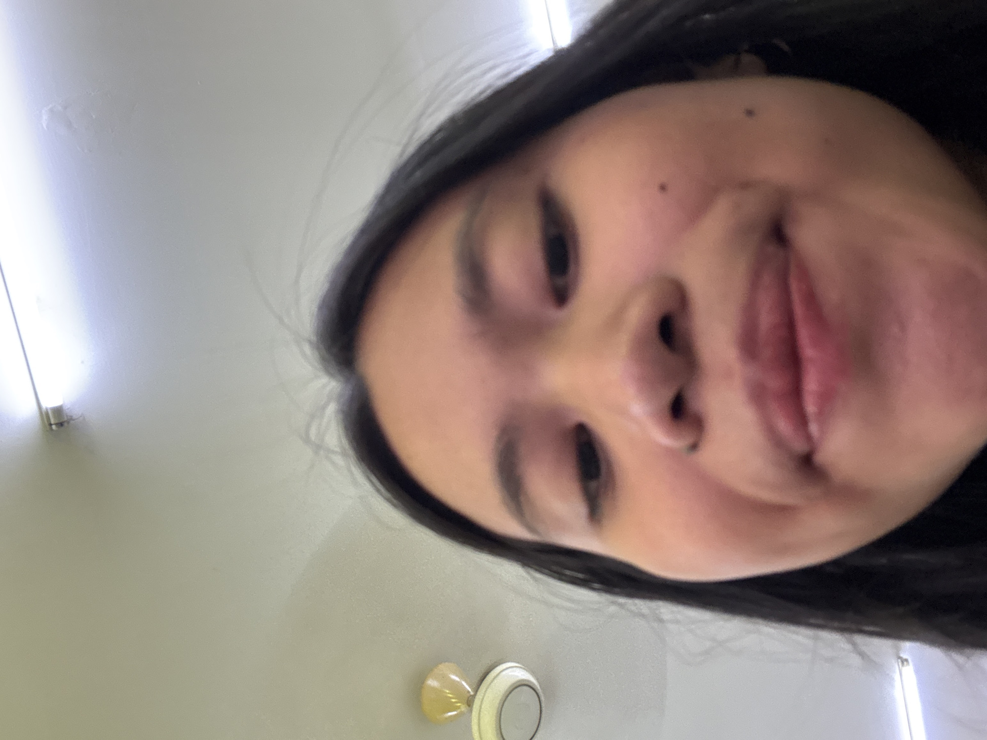 A close-up selfie taken from a low angle shows a young Asian woman's face and upper body. She has long, dark hair framing her face. Her eyes are open, and she appears to be looking upwards towards the camera. Her lips are slightly parted, and she has a mole on her right cheek. The background is a ceiling with a white fluorescent light fixture and a ceiling fan. The environment appears to be indoors, possibly a room or a hallway in East Jakarta, Indonesia, given the context of the image. The overall lighting is bright, suggesting artificial light is the primary source.