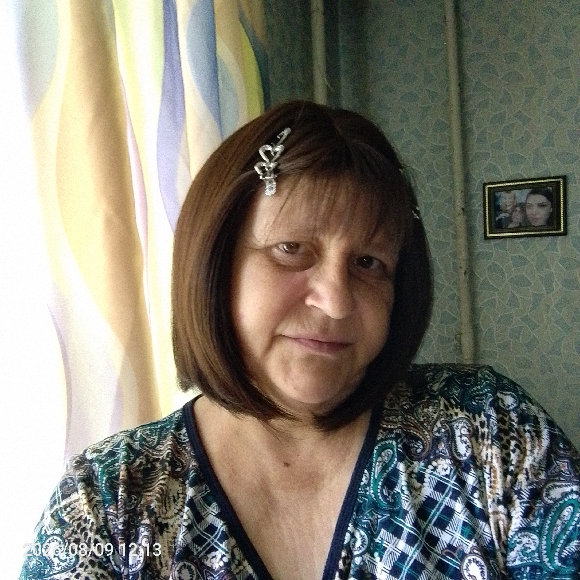 A woman with shoulder-length brown hair and a decorative hair clip is seen from the chest up, looking towards the camera. She is wearing a dark top with a paisley and plaid pattern in green, white, and blue. The background consists of a light-colored curtain on the left and a patterned wallpaper on the right, with a framed photograph hanging on the wall behind her. The photograph appears to contain three people. A date stamp "2016/08/09 12:13" is visible in the lower left corner of the image.
