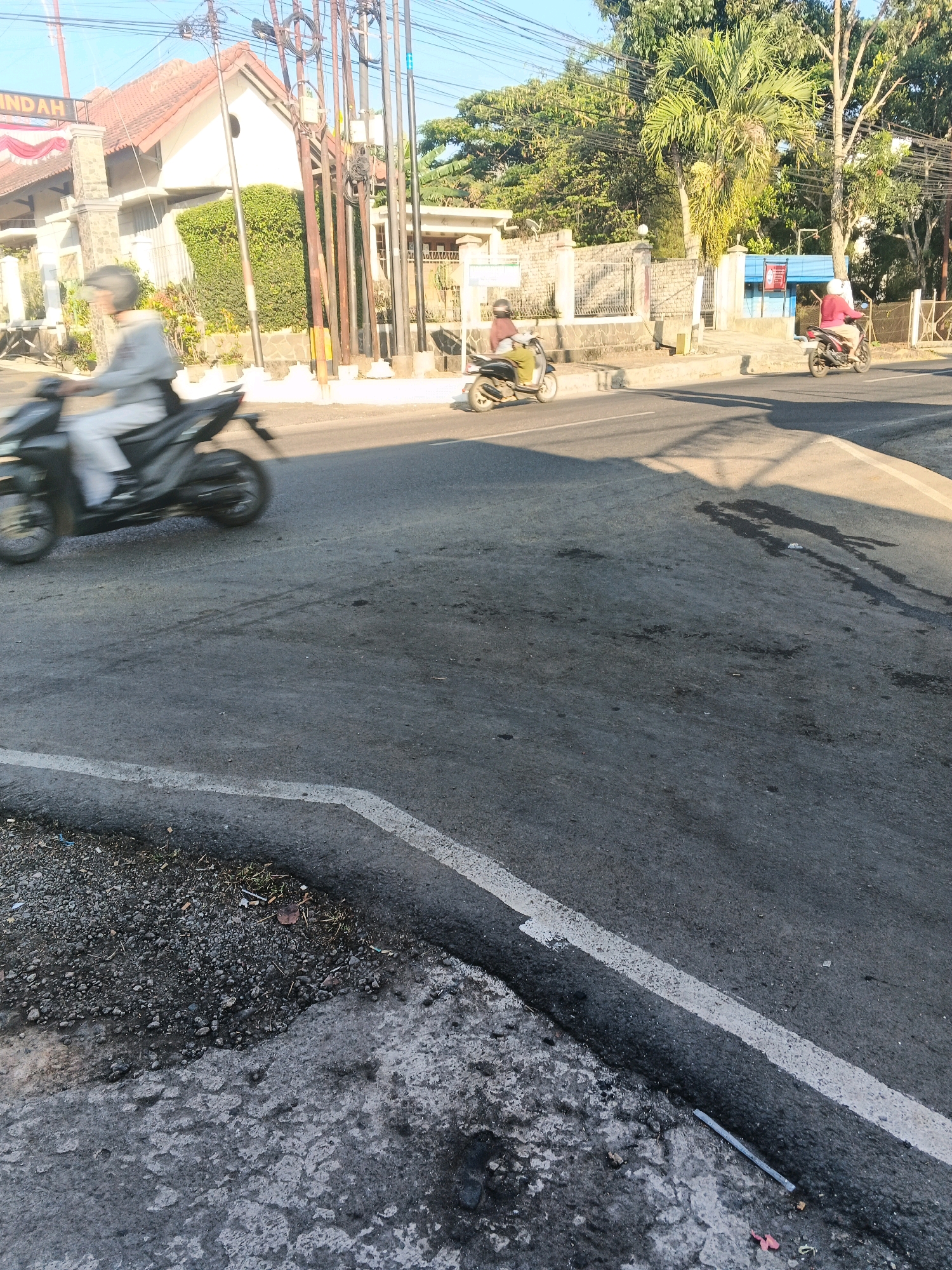 A street scene in Semarang, Indonesia, features a light gray motorcycle moving from left to right in the foreground. The rider, wearing a helmet and light-colored clothing, is blurred due to motion. In the middle ground, another motorcycle with a rider in a light brown shirt is also moving from left to right, also blurred. To the right, a third motorcycle with a rider in a red shirt moves in the same direction. The road surface is asphalt with visible tire marks and some dark stains, possibly oil or water. On the left side of the road, there is a gray curb with gravel and some vegetation. In the background, there are buildings, trees, and utility poles with numerous wires. The sky is clear and blue.