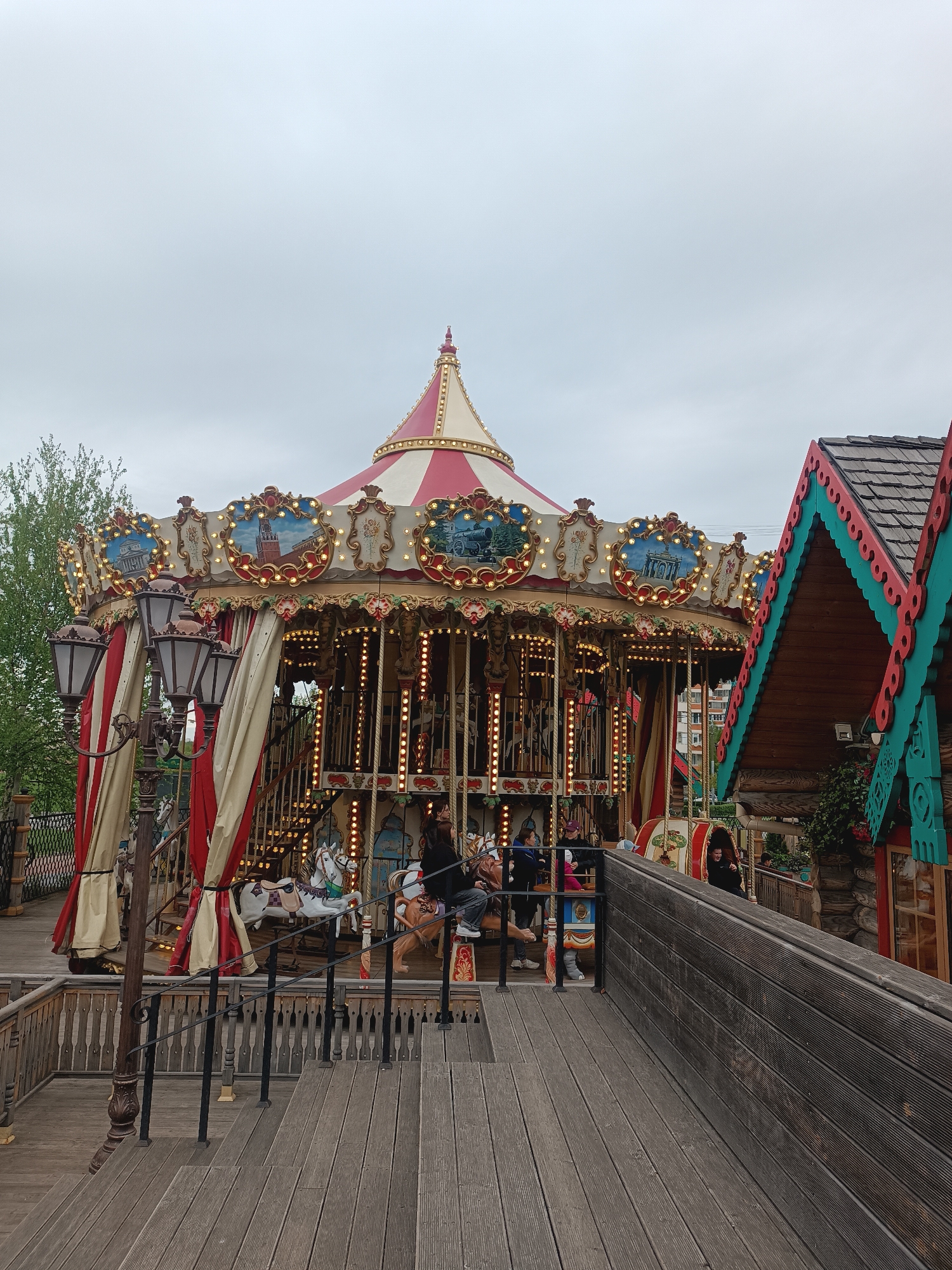 The image captures a carousel ride in Moscow, Russia. The carousel is adorned with red and white striped fabric and illuminated with light bulbs, creating a festive atmosphere. The top of the carousel features paintings of Moscow landmarks. People are seen riding the carousel. Adjacent to the carousel is a wooden structure with ornate detailing and a colorful roof, likely a part of the amusement park or fair setting. The scene is set on a wooden deck with railings and stairs, leading to the carousel. The sky is overcast, creating a soft, diffused light.