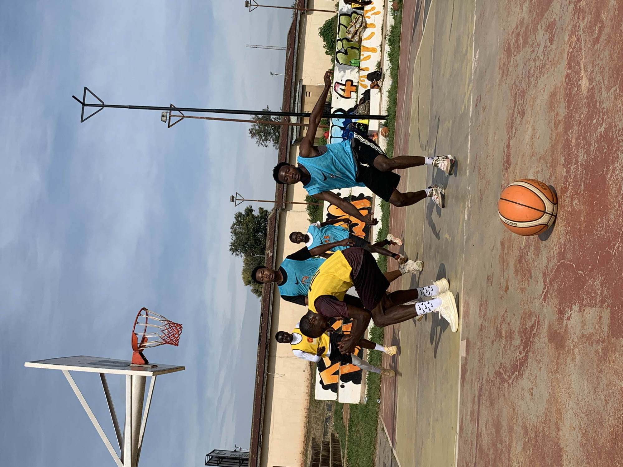 A group of young men are playing basketball on an outdoor court in Gura Top, Nigeria. The court is made of concrete and has painted lines. There is a basketball hoop with a red net. The men are wearing athletic clothing, including jerseys and shorts. One man in a maroon and yellow jersey is crouched low, appearing to be defending. Another man in a blue jersey is in a defensive stance. A basketball lies on the court in the foreground. In the background, there is a building with graffiti on its wall and trees. The sky is partly cloudy.