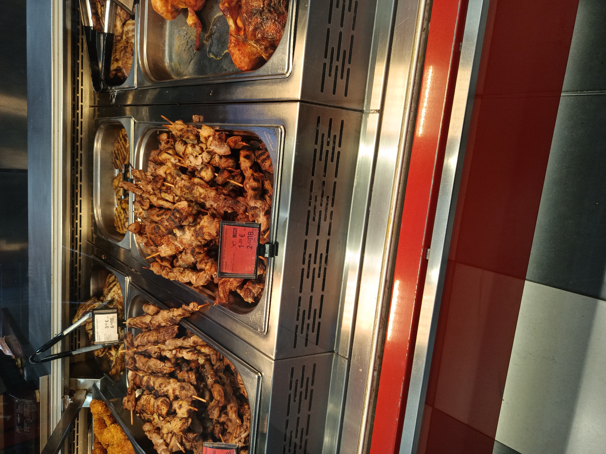 The image shows a display case filled with grilled meat skewers, likely in a grocery store or deli in Sofia, Bulgaria. The skewers are arranged in metal trays on multiple levels. The display case has a metallic exterior with ventilation slots. One sign displays a price of 2.52лв, the currency of Bulgaria. Tongs are available for customers.
