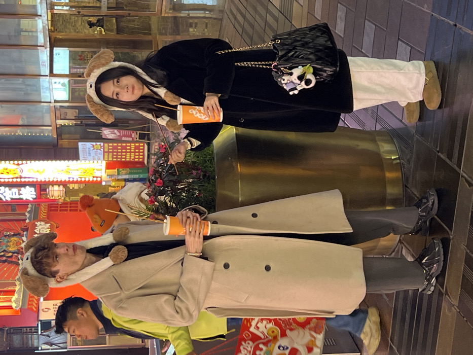 The image displays two individuals, an adult male and an adult female, standing on a tiled pavement at night in a commercial area of Yuzhong District, China. Both are wearing matching white and brown plush hats with floppy ears and holding orange and white beverage cups with straws.
The male, positioned on the left, has light brown hair and a fair complexion. He wears a beige double-breasted trench coat over a black top and dark grey trousers, paired with dark, reflective slip-on shoes. A bracelet is visible on his left wrist. His expression is neutral.
The female, on the right, has long dark hair and an Asian ethnicity. She wears a black faux fur coat over white wide-leg trousers and tan closed-toe slip-on shoes. In her left hand, she holds a wooden skewer, appearing to consume street food, while holding her beverage cup in her right hand. She carries a black quilted handbag with a gold chain strap, adorned with white and green animal-shaped keychains. Her expression is slightly pensive.
Between the two subjects is a large, reflective gold cylindrical planter containing green foliage with small red flowers. The background features brightly illuminated commercial buildings with numerous vertical neon signs displaying Chinese characters in red, yellow, and orange hues. Partial views of other individuals, including a man in a lime green jacket and another in an orange beanie, are visible. The overall setting is an artificially lit urban street or marketplace.
