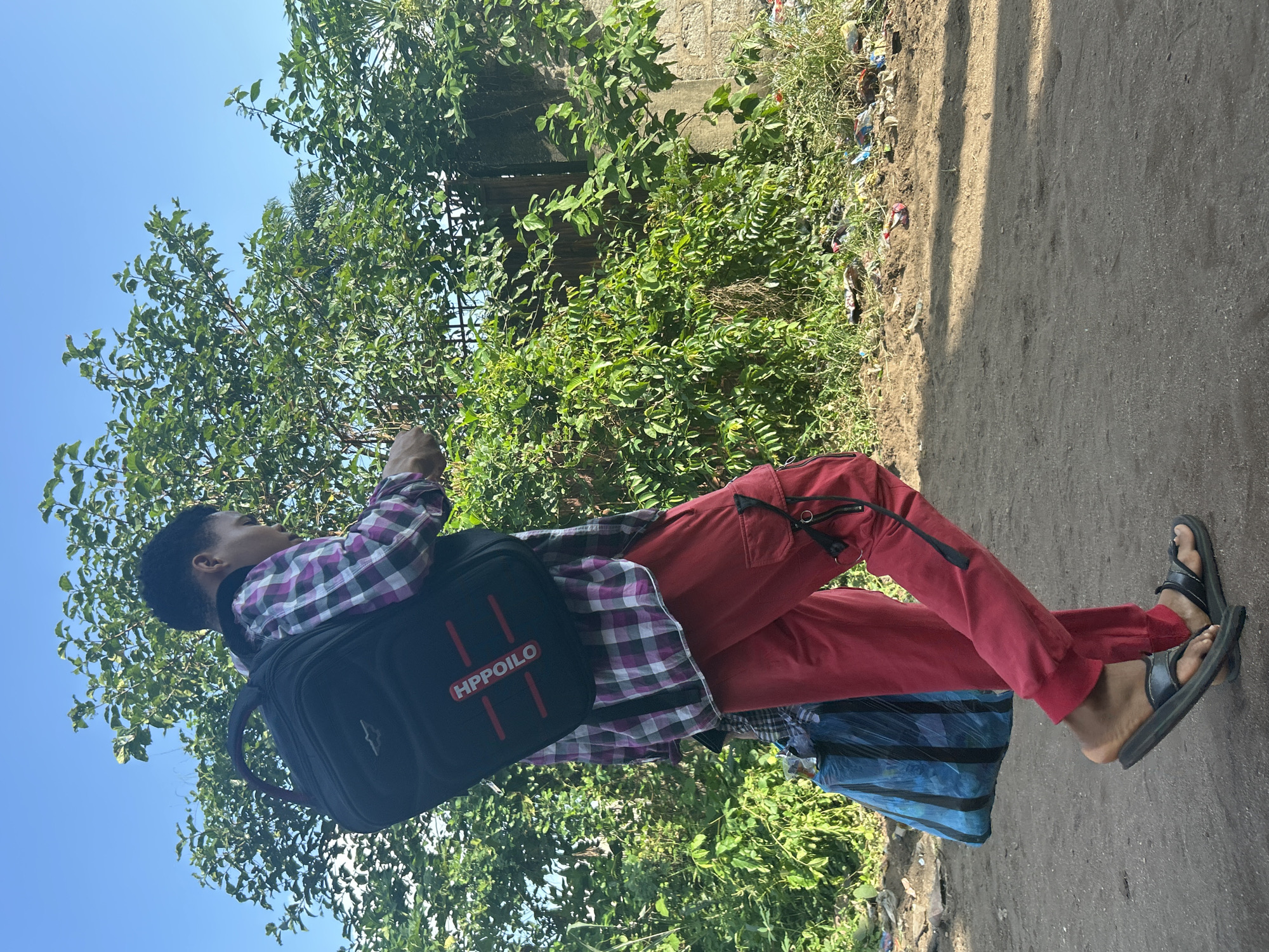 A young Black person with short dark hair walks from left to right across a dusty, unpaved path in Ihiagwa, Nigeria, under a clear blue sky. The person is wearing a purple and white plaid shirt over a dark vest, and bright red cargo pants with black accents. A black backpack with "HPPOILO" in red lettering is worn on their back, and a blue and black striped bag is carried in their left arm. Black flip-flops are on their feet. Lush green trees and foliage line the path, with a stone wall and metal gate visible in the background. The ground is littered with various debris, suggesting an outdoor, possibly semi-rural or urban fringe environment. The person appears to be looking down at their phone.
