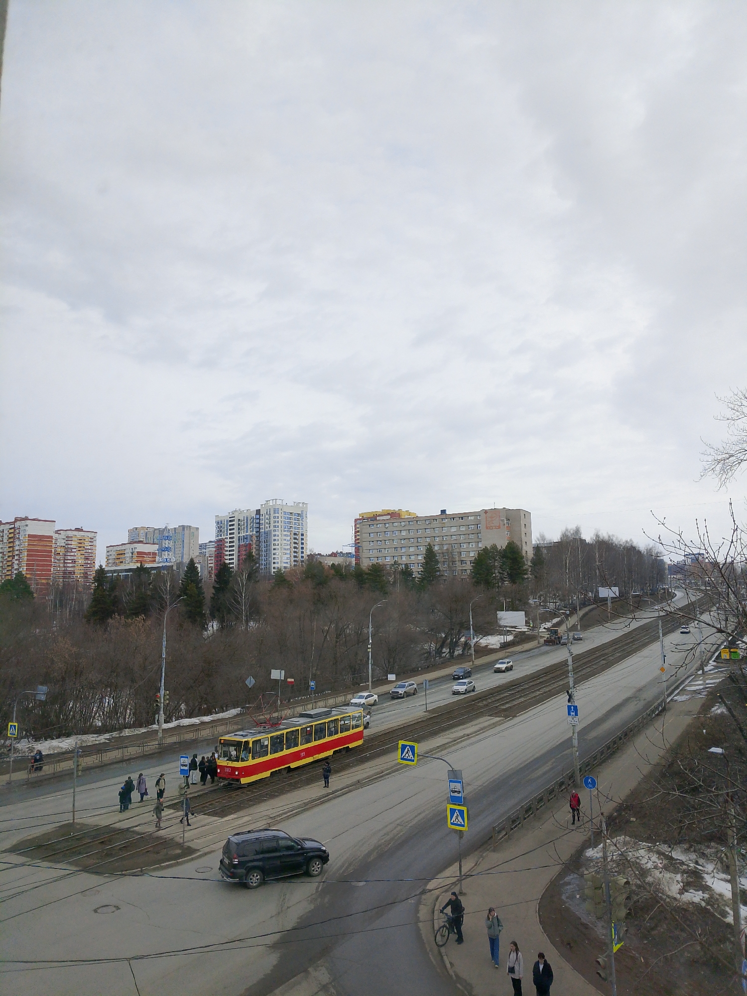 The image shows a street scene in Izhevsk, Russia, under a cloudy sky. In the foreground, there is a multi-lane road intersection with a yellow and red tram stopped at a pedestrian crossing. People are crossing the street near the tram, and a black SUV is positioned on the road nearby. Pedestrians are also walking along the sidewalks on either side of the road. Further back, a multi-lane road stretches into the distance with other cars driving on it. Buildings of varying heights and colors are visible in the background beyond the road. There are bare trees and patches of snow alongside the road and in the background, suggesting a cold time of year. Streetlights and pedestrian crossing signs are also visible along the road.