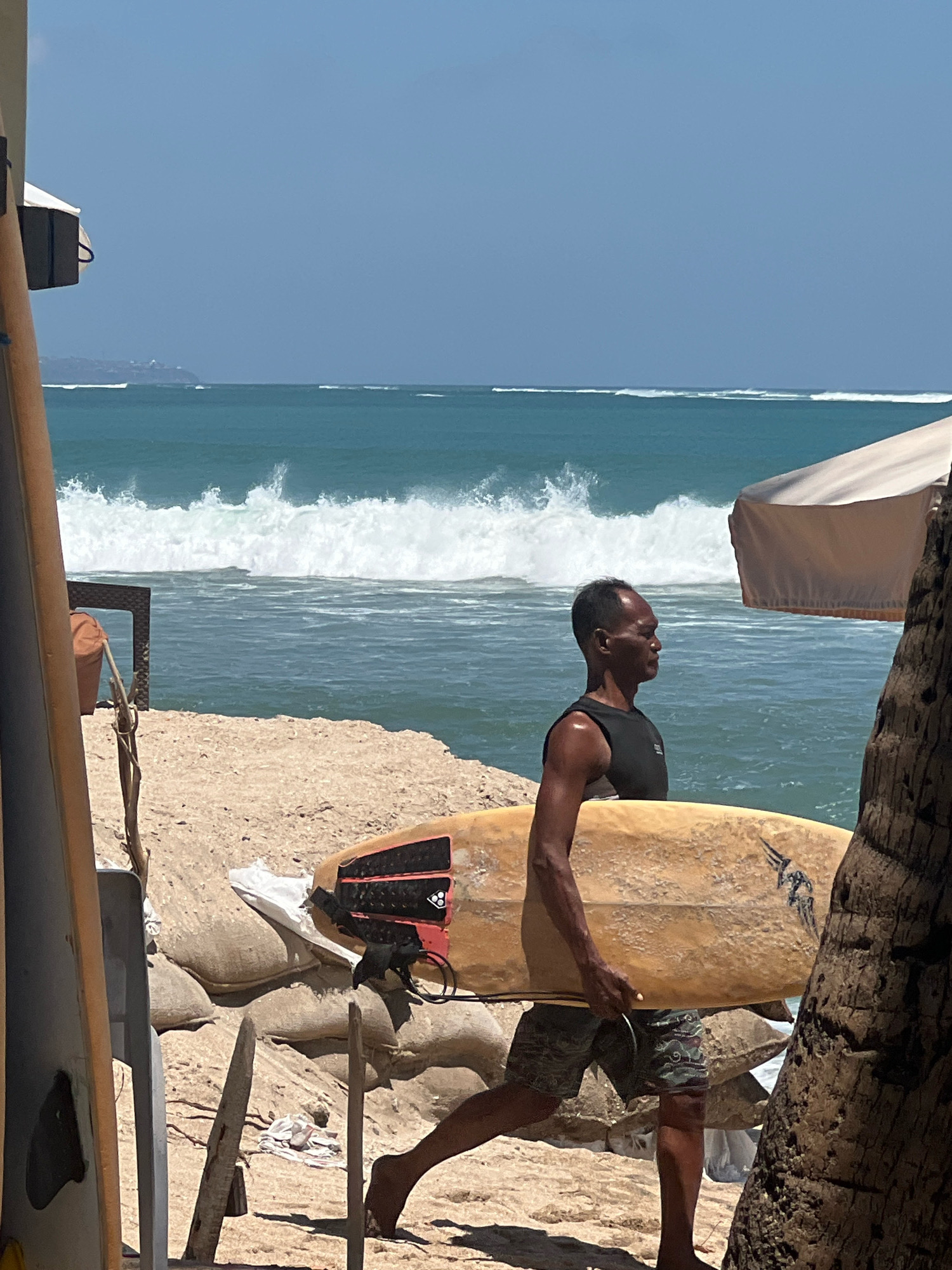 A medium shot captures a man walking along the beach in Kuta, Indonesia, carrying a surfboard. He is wearing a black tank top and camouflage shorts, and his bare feet are on the sandy ground. The surfboard, a pale yellow color, has a black leash attached. In the background, the ocean displays breaking waves and a clear blue sky. To the left of the man, sandbags are stacked. Further in the background, white tents and palm trees are visible, hinting at a beachside establishment. The overall scene suggests a typical day at a popular Indonesian surf spot.