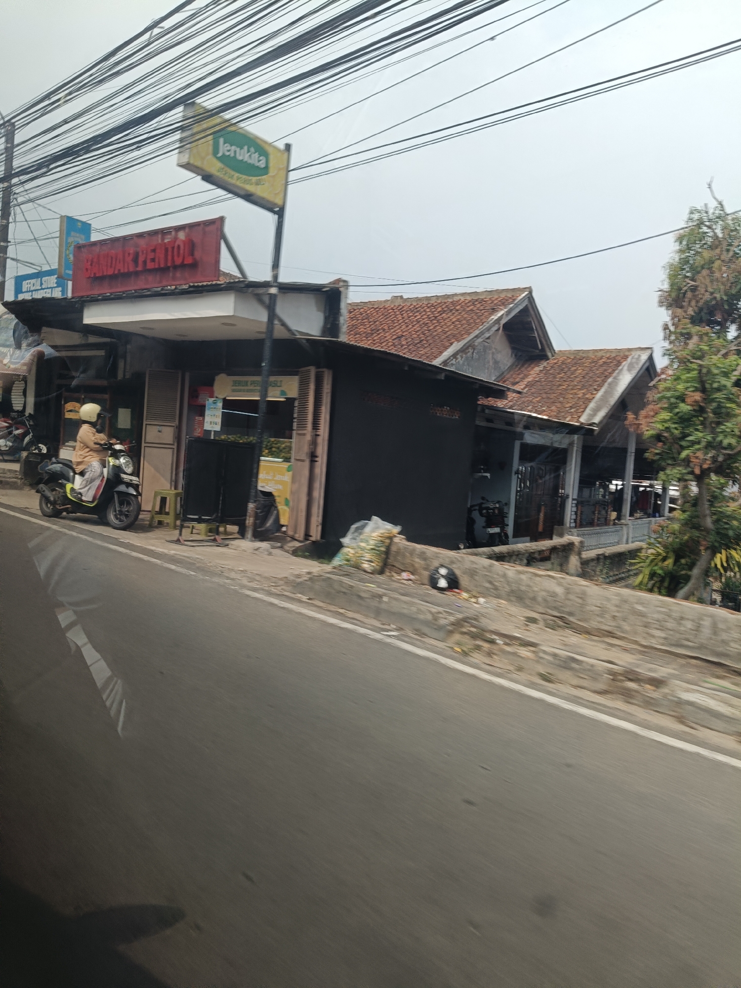The image depicts a street scene in Pandeglang, Indonesia, on a cloudy day. A prominent red sign with white lettering reading "BANDAR PENTOL" is mounted above the entrance of a building, suggesting a food establishment. To the left of this sign, a yellow sign with green lettering reads "Jerukita," likely indicating a brand of citrus drink. Below the "BANDAR PENTOL" sign, a white awning extends over the building's facade.
In front of the building, a person wearing a light brown long-sleeved shirt and white pants, with a yellow helmet, is seated on a black and white scooter. The scooter is parked on the sidewalk. Adjacent to the scooter, a small green stool is placed on the ground. The building's storefront appears to have open windows or doors, revealing products on shelves within, including what appear to be fruits, possibly oranges, displayed behind a counter. Several trash bags are visible near the storefront.
To the left of the building, a smaller blue sign is partially visible, and to the right, trees and parts of other buildings with tiled roofs are present. The foreground is dominated by a road, with the reflection of structures and power lines visible on its surface, indicating movement through the scene. The environment suggests a commercial area along a roadside.