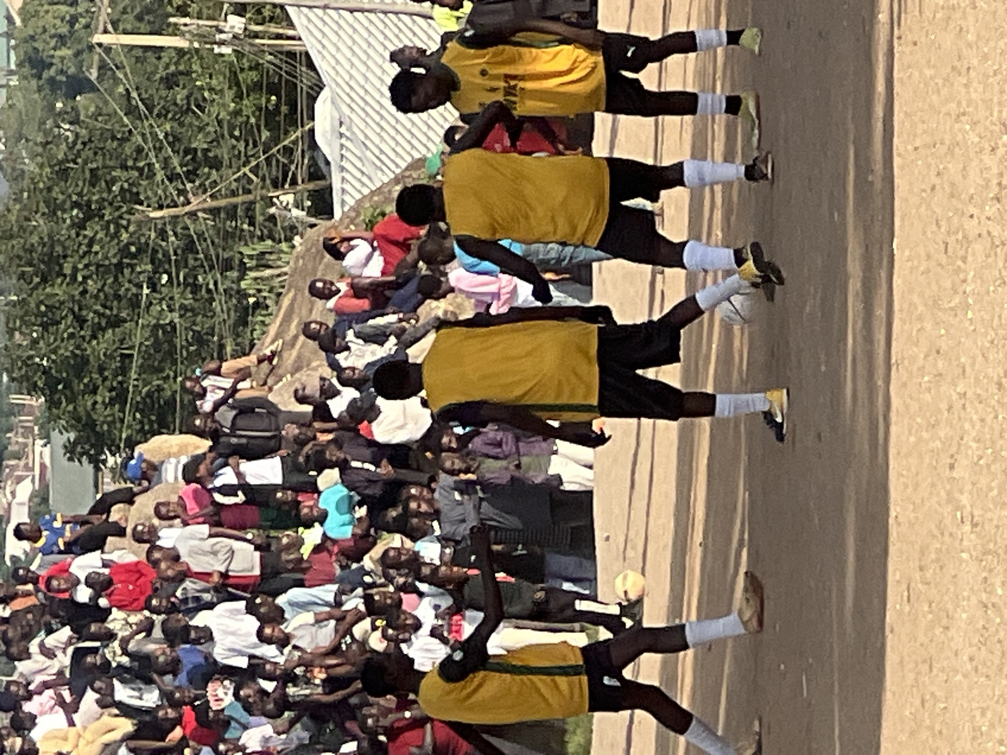 The image depicts a street soccer match taking place in the city of Jos, Nigeria, with a crowd of spectators gathered on a hillside overlooking the field. The players, dressed in yellow jerseys and black shorts, are actively engaged in the game, with one player in the foreground preparing to kick a soccer ball. The spectators, a diverse group of men, women, and children, are seated or standing on the rocky terrain, their attention focused on the match. The environment is dusty and arid, typical of the Jos Plateau. Buildings and trees are visible in the background, suggesting a semi-urban setting. The overall scene is one of lively community engagement and sporting activity.