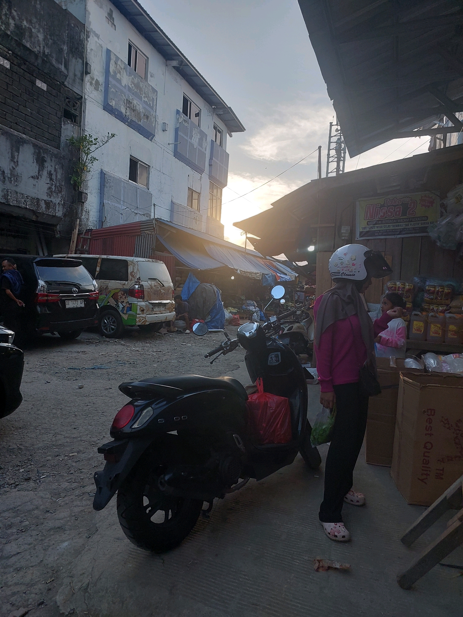 The image depicts an outdoor market scene, likely in Ambon, Indonesia, with a mix of buildings and stalls. In the foreground, a black scooter is parked on a dirt and concrete ground. To the right of the scooter, a woman wearing a pink long-sleeved shirt, a grey headscarf, and black pants stands with a white helmet on her head. She is holding a plastic bag filled with green items. Next to her, a young girl in a pink long-sleeved shirt and white pants is visible. Behind them, a stall with yellow containers of liquid and bags of goods is set up. A large cardboard box with the text "Best Quality" is on the right side of the frame. In the background, two vans are parked. One is black with a license plate that reads "DE 1472 AP," and the other is white with a colorful cartoon decal. A building with peeling paint and blue accents is on the left. The sky is visible above, with some clouds and the setting sun casting a warm glow.