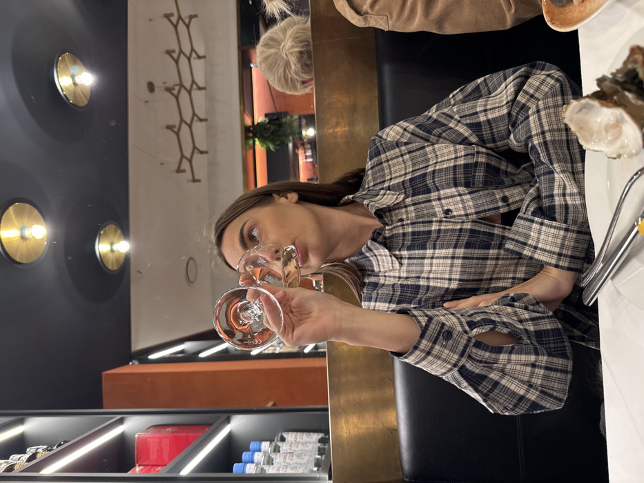 The image depicts an indoor scene at a restaurant or bar located in Verona, Italy. A young adult female with light complexion and brown hair, wearing a navy and beige plaid button-down shirt, is seated at a dark booth. She holds a stemmed glass containing a clear liquid to her lips, appearing to drink or prepare to drink. Her gaze is directed upward and to the right.
On the white table in the foreground, a white plate holds an opened oyster and a set of silver-colored cutlery (fork and knife). To the right of the main subject, a person's arm in a tan jacket is partially visible. In the background, behind a brass-colored partition, another person with blonde hair tied in a bun is partially visible. Further back, a bar or counter area is present, featuring a potted plant, bottles, and warm orange lighting. The ceiling is dark, featuring multiple circular recessed light fixtures with exposed bulbs, and a large, geometric, brass-toned decorative light fixture. To the left, a dark shelving unit with internal backlighting displays various bottles and red-labeled items.