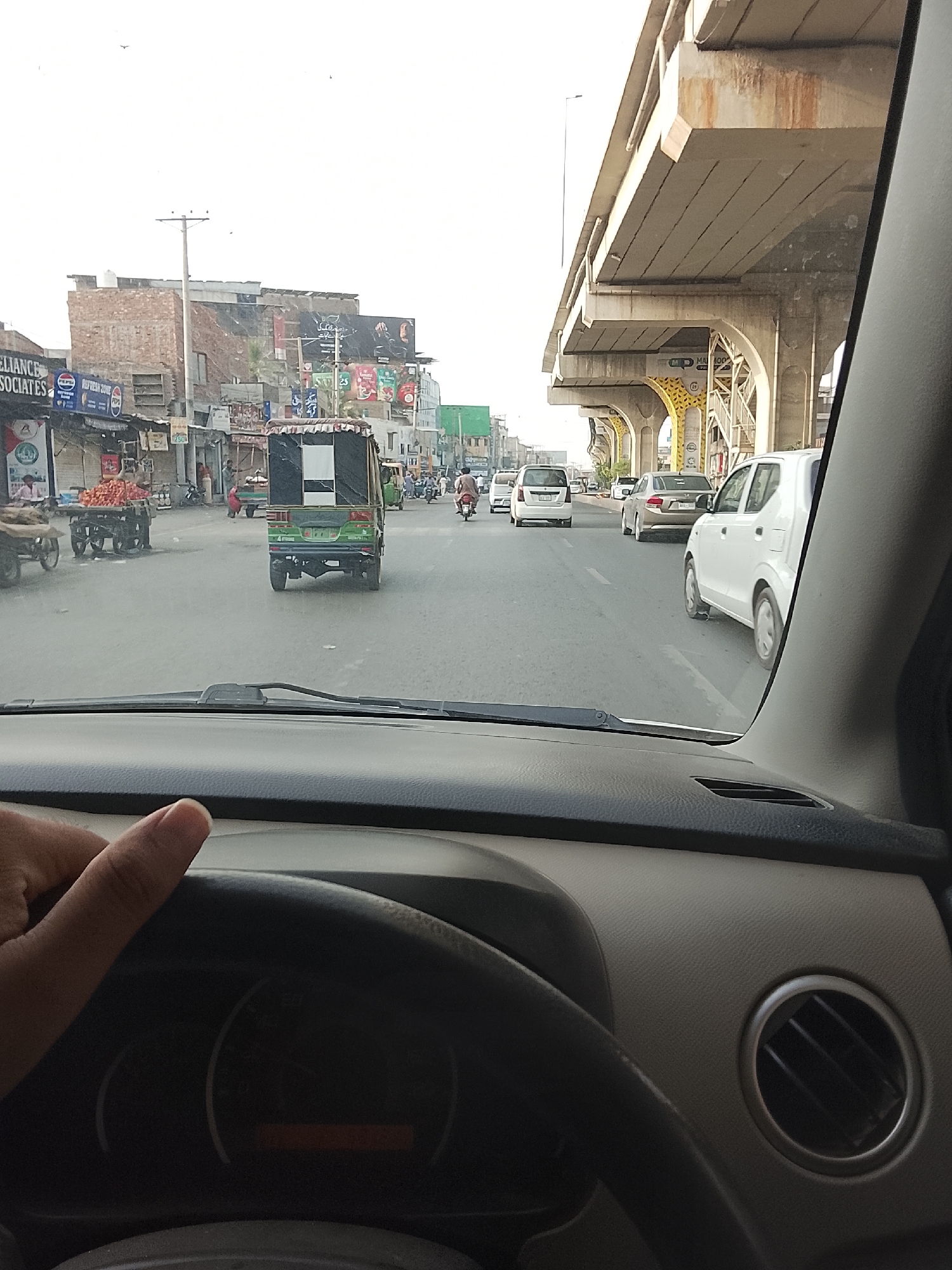 The image shows a view from inside a car in what appears to be Gulgasht Colony, Pakistan, indicated by signs in the background, urban structures, and vehicles on the road. The driver's hand is visible on the steering wheel. In front of the car are an auto-rickshaw and other vehicles, including cars and motorcycles. The road leads through a cityscape with buildings on either side. An elevated road or bridge is on the right side of the image. There are people visible on the sides of the road, some walking, some on carts.