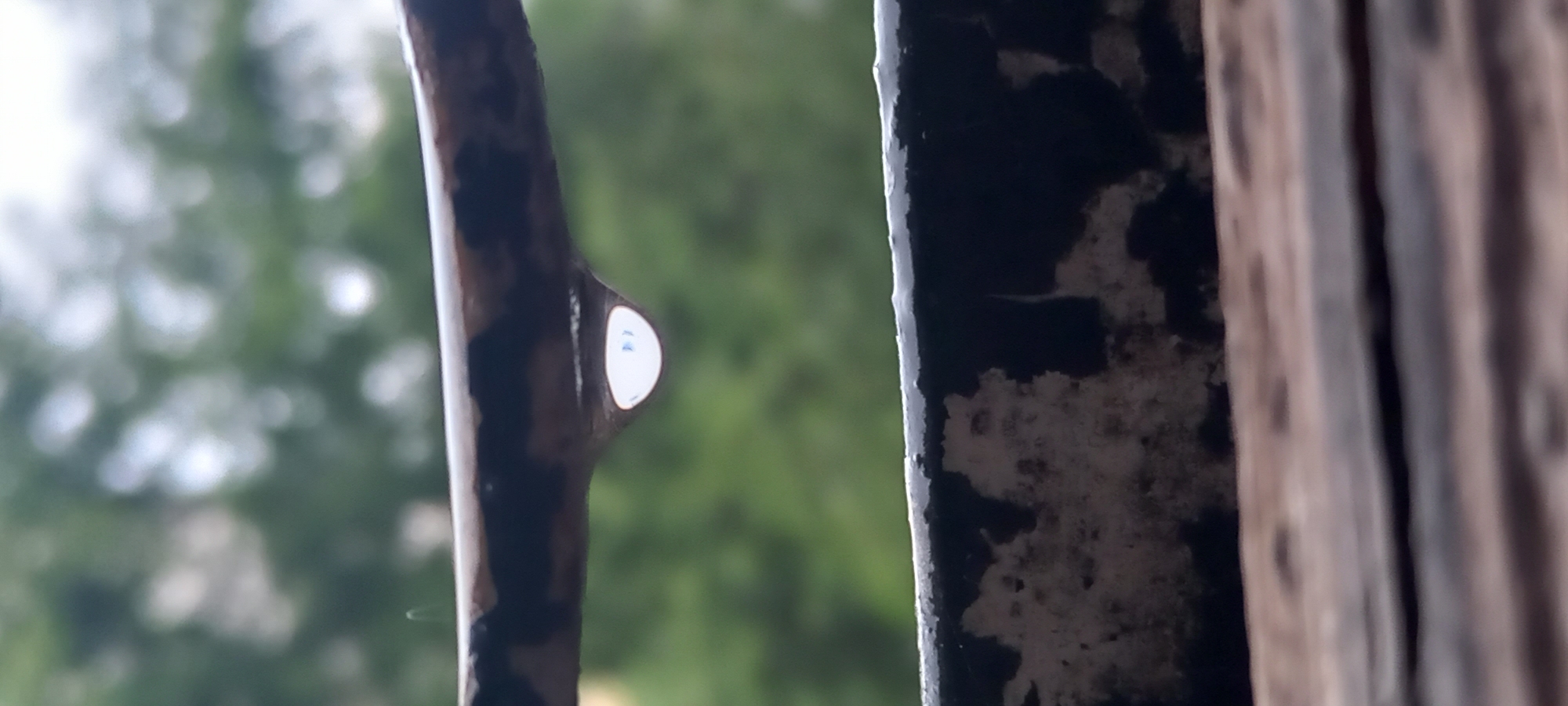 A close-up, vertical shot captures a single water droplet suspended from a dark, horizontal bar. The droplet is teardrop-shaped and appears clear, reflecting a bright, white, slightly bluish light. The bar it hangs from is textured and weathered, with patches of rust or peeling paint. Below this bar is another horizontal surface, darker and also appearing weathered or textured. In the background, the scene is heavily blurred, suggesting out-of-focus greenery and a bright sky, indicative of an outdoor setting. The image appears to be taken at eye level. There are no people or any indication of a specific location within Kryvyi Rih, Ukraine.