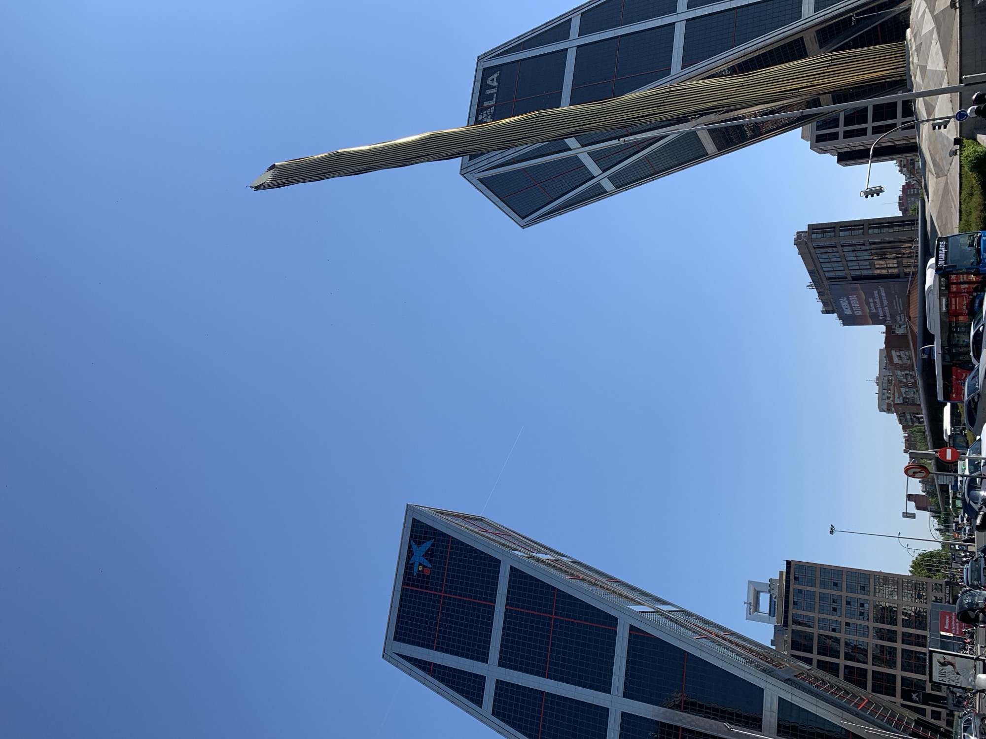 The image shows the KIO Towers in Madrid, Spain, with a clear blue sky. The two iconic inclined skyscrapers are the main subject, with the left tower featuring a blue star logo. A golden, ribbed spire rises between the two buildings. The foreground shows a busy street with cars, buses, and traffic signals, indicating an urban environment. Several other buildings and trees are visible in the background, contributing to the city landscape. The sunlight is bright, casting shadows and highlighting the architectural details of the towers.
