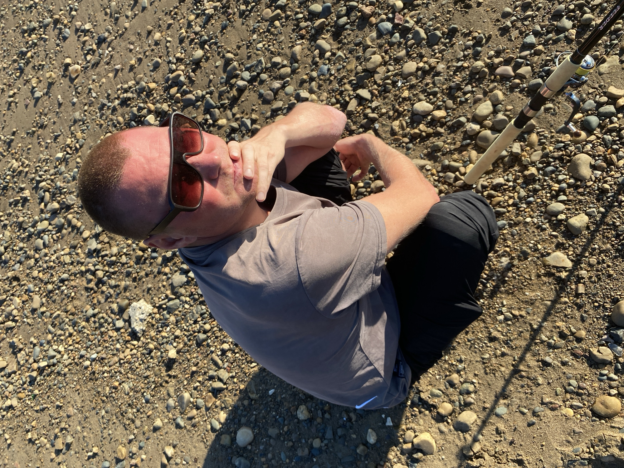 A man with fair skin, a shaved head, and sunglasses is squatting on a pebble-covered ground. He is wearing a grey t-shirt and black pants. His left hand is on his chin, and his right arm is bent. A fishing rod is on the ground next to him, with the handle facing the camera. The background is a vast expanse of pebbles. The image does not provide any information about the city of Chaikovsky, Russia, or any specific location within it.