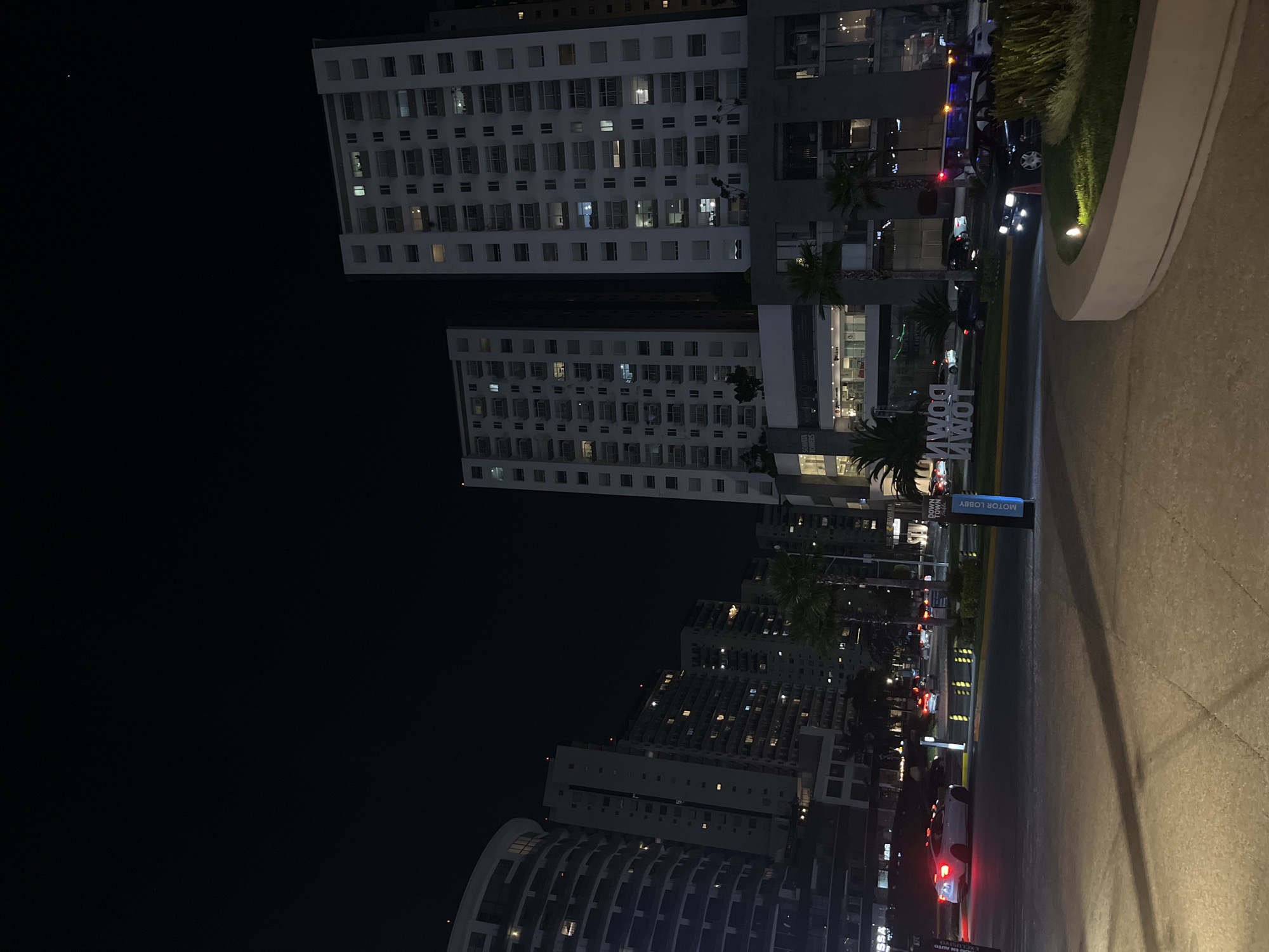 The image shows an urban nighttime scene in San Bernardino Tlaxcalancingo, Mexico. Tall, lit-up residential buildings dominate the background. A road runs through the middle ground with parked cars and moving vehicles, including one with red taillights. A sign reading "DOWN TOWN" and "MOTOR LOBBY" is visible next to the road. The foreground consists of a paved surface. No people are visibly identifiable.