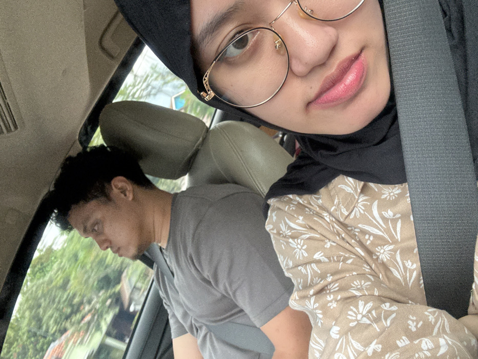 Inside a car, a woman wearing a black hijab and gold-rimmed glasses poses for a selfie. Her face is partially framed by the car's seatbelt. She wears a beige patterned blouse. Seated in the back, a man in a gray t-shirt looks down. Greenery is visible through the car's window. The car's interior is mostly shades of gray and beige.
