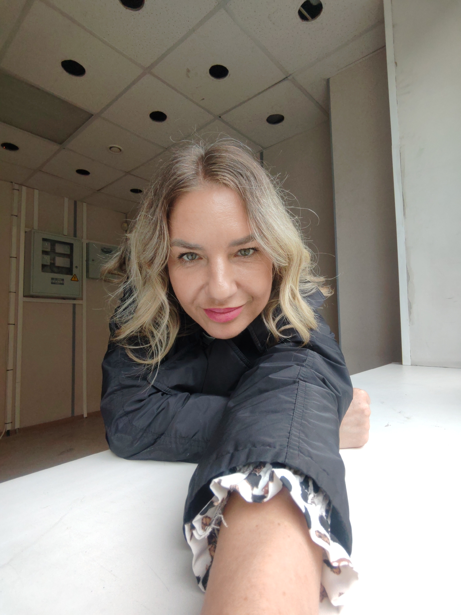 A selfie of a woman with blonde hair, wearing a black jacket with a patterned white and black cuff, taken in an indoor setting. The woman has green eyes and is wearing pink lipstick. The background features a tiled ceiling with recessed lights and beige walls with electrical boxes. The photograph was likely taken in an industrial or commercial building. There is no evidence in the image to suggest this photo was taken in Petropavlovsk-Kamchatsky.