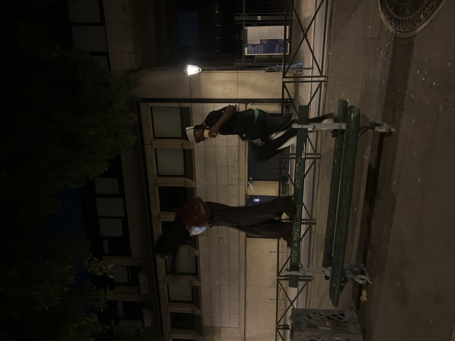 The image captures a nighttime urban scene in Paris, France. Two individuals are depicted in dynamic motion on and around a green public park bench, characteristic of Parisian street furniture with its ornate dark metal supports and wooden slats.
On the left, one individual, obscured by motion blur, stands atop the backrest of the bench, appearing to traverse it. They are dressed in dark, loose-fitting clothing and carry a reddish-brown item. On the right, a second individual is captured mid-action, with their right leg raised, stepping or vaulting over the bench's backrest. This person wears a dark short-sleeved shirt, dark trousers with a white stripe, and a white headband, holding a light-colored cup to their mouth. An earring is visible on their left ear.
In the mid-ground, a multi-story building with a light-colored stone facade and numerous rectangular windows extends across the frame. To the right, a traditional black Parisian street lamp with a white globe illuminates the area. A black metal fence or barrier is situated behind the bench. To the left of the bench, a decorative metal trash receptacle is present. The foreground consists of a dark paved surface, with a circular metal manhole cover partially visible on the far right. Dark tree foliage is visible against the dark night sky in the upper portion of the image. Graffiti, including the words "KAYE" and "NIKE," is visible on the backrest of the bench.