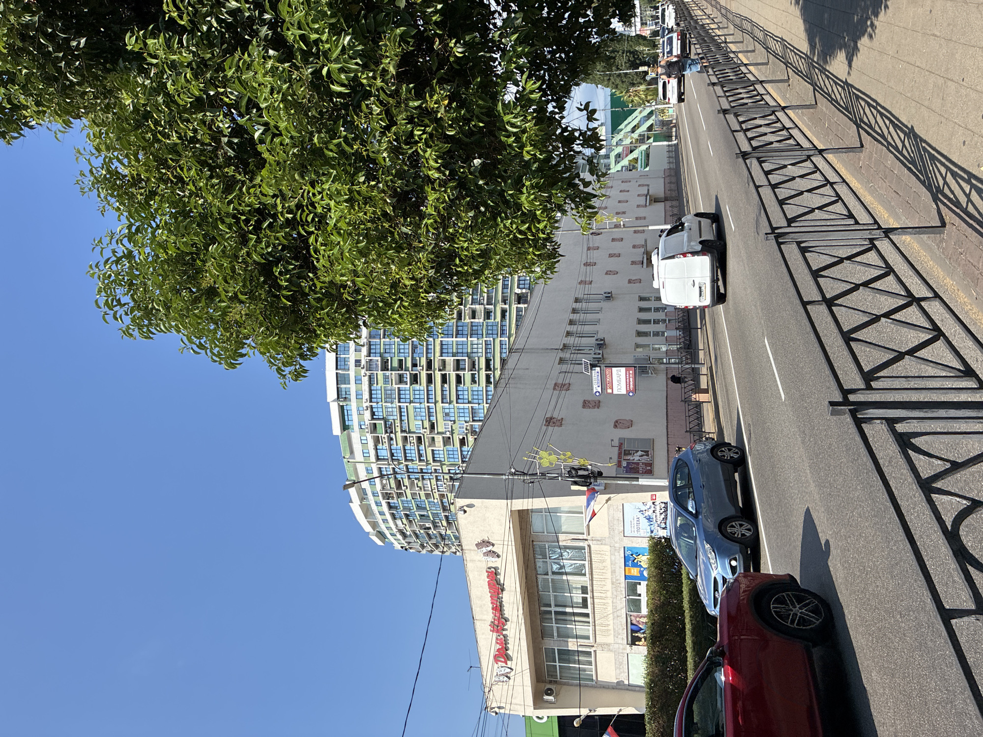 The image depicts a street scene in Sochi, Russia, on a sunny day. In the foreground, a black metal fence lines the sidewalk, casting long shadows across the pavement. A red SUV is partially visible on the left, and a blue hatchback is driving in the lane next to it. Beyond the cars, a white van is seen driving down the road.
On the right side of the street, a building with a sign in Russian that reads "Дом Культуры" (House of Culture) is prominent. Further down the street, a tall, modern building with a curved facade and numerous balconies is visible against a clear blue sky. A large, leafy green tree branches out from the right side of the frame, partially obscuring the sky and the taller building.
Further down the street, a few pedestrians are walking, and several other vehicles are present. The environment suggests a relatively busy urban area with a mix of older and newer architecture.
