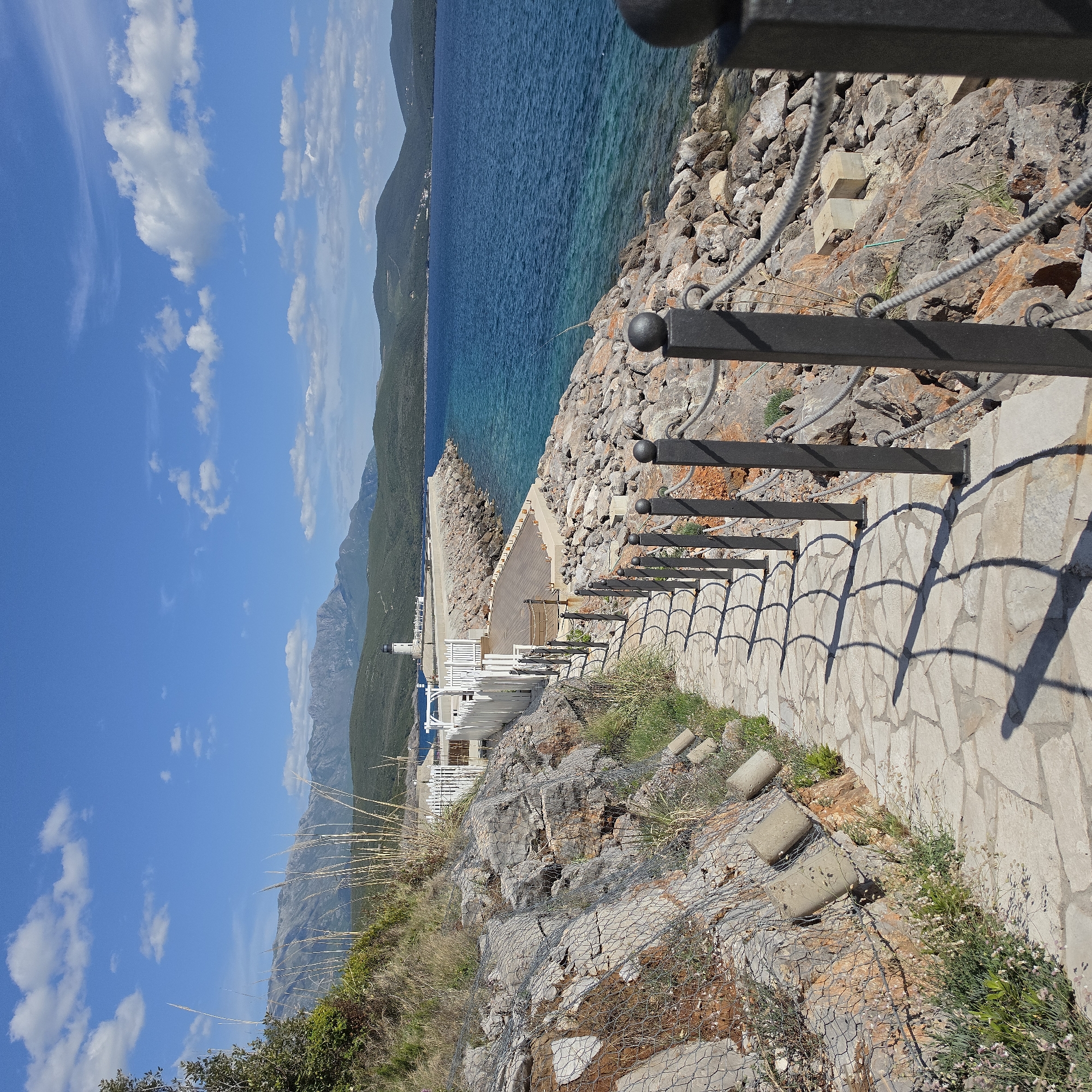 The image displays a serene coastal landscape, likely in Radovići, Montenegro, under a bright, sunny sky. The scene is viewed from a paved stone pathway winding down a cliffside towards the sea.
In the foreground, a path constructed from light-colored, irregularly shaped flagstones descends. A black metal railing with spherical finials borders the path, casting prominent, arching shadows onto a contiguous stone wall. The rocky cliff face adjacent to the path features reddish-brown and grey stones, interspersed with dry grasses and small plants. Sections of the cliff are covered with wire mesh, suggesting rockfall mitigation. Ropes with metal rings are also visible, acting as informal barriers.
Further down, closer to the water, several white buildings, possibly part of a resort or eatery, are visible with white railings or fences. A substantial concrete pier or jetty extends into the water from this area, with a small lighthouse or beacon situated at its far end.
The water of the sea transitions from a clear turquoise near the shore to a darker, deep blue further offshore. In the background, a range of verdant, rugged mountains stretches across the horizon, complementing the expansive blue sky dotted with fluffy white clouds.
The time of day appears to be mid-morning or early afternoon, indicated by the strong shadows. The weather is clear and sunny. There are no people visible, and no specific activities are occurring, lending a tranquil and picturesque atmosphere to the scene. No visible text is present in the image.