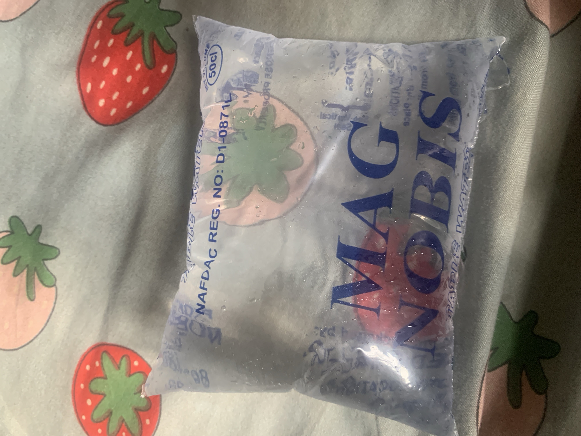 A close-up, eye-level shot shows a sealed plastic sachet of water on a light blue fabric with strawberry patterns. The sachet is transparent and filled with water, with small droplets visible on the exterior. The brand name "MAG NOBIS" is printed in large, dark blue capital letters on the sachet. Above the brand name, "NAFDAC REG. NO: D1-08710" and "NET VOLUME 50cl" are printed. There is a stylized image of a strawberry on the sachet as well. The sachet appears to be lying on a bed or similar surface. The image context indicates the location is Jalingo, Nigeria. There are no people or discernible actions in the image.