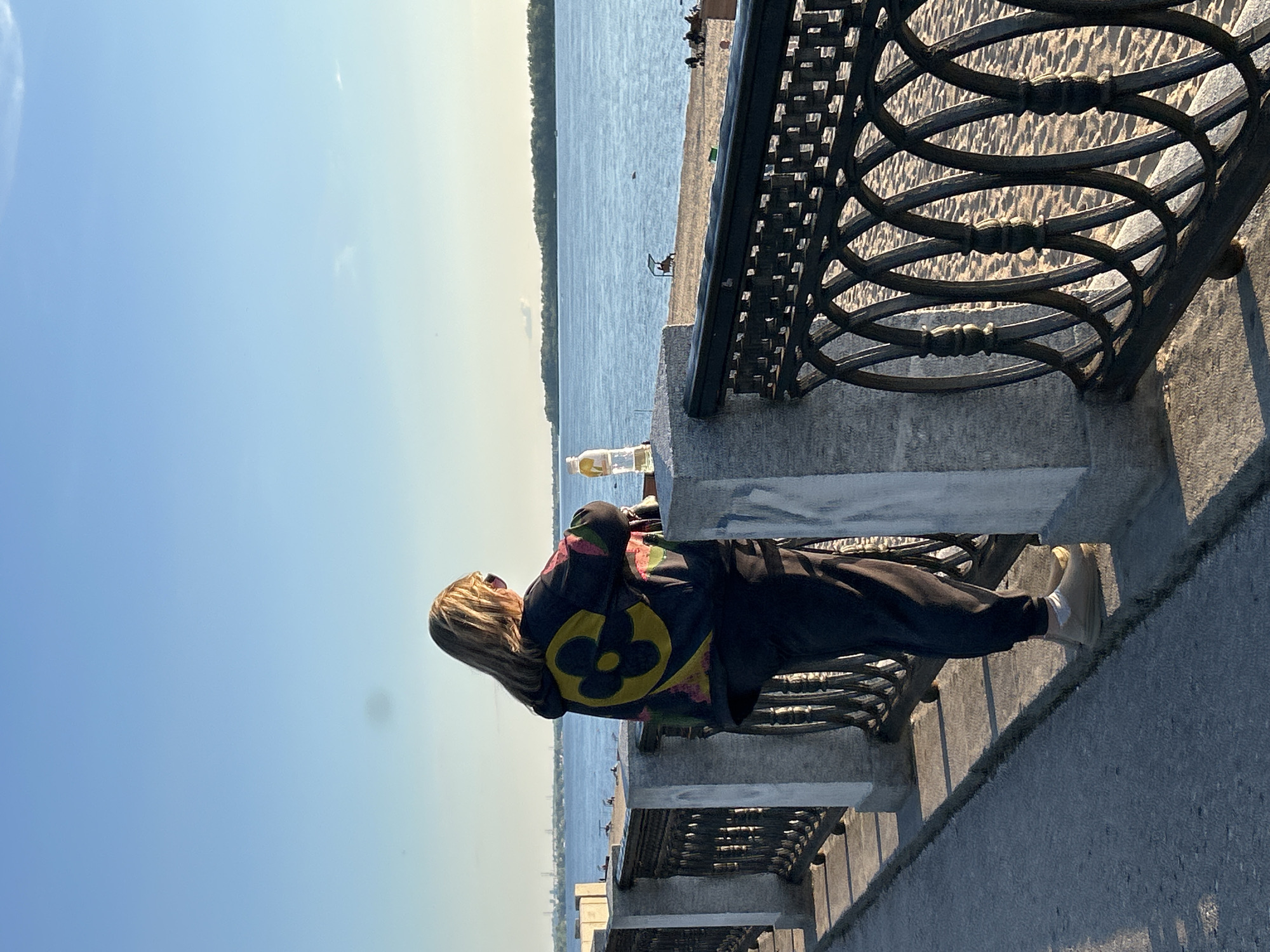 The image shows a person from the back, standing on a paved area next to a body of water. The person has long, blonde hair and is wearing a dark jacket with a colorful, circular emblem on the back, and dark pants. They are facing away from the camera, looking out at the water. To the right of the person is a decorative metal railing, and beyond that, a sandy area. The water stretches to the horizon, where a tree-lined shore is visible. The sky is a clear blue, with the sun casting long shadows, suggesting it is late afternoon or early evening. The setting appears to be a waterfront promenade or park. No visible text is present in the image. The overall mood is calm and serene, with the person appearing contemplative or relaxed.