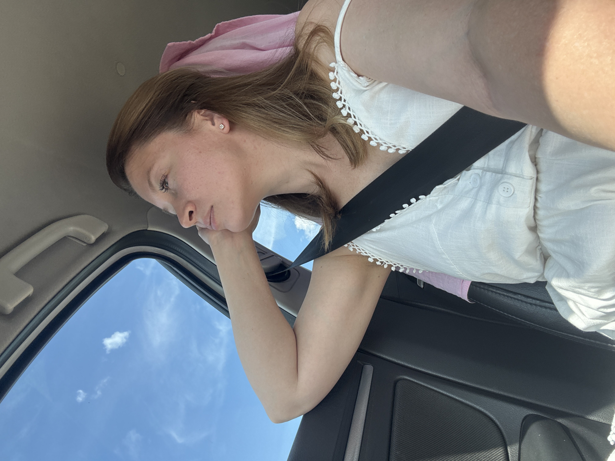 A woman is sitting in the passenger seat of a car, looking out the window. She has light brown hair and is wearing a white dress. A pink towel is draped over the back of the seat behind her. The car is in motion, as indicated by the blurred background and the sky visible through the open sunroof. The scene appears to be a sunny day with a few clouds. The location is not specified in the image or context.