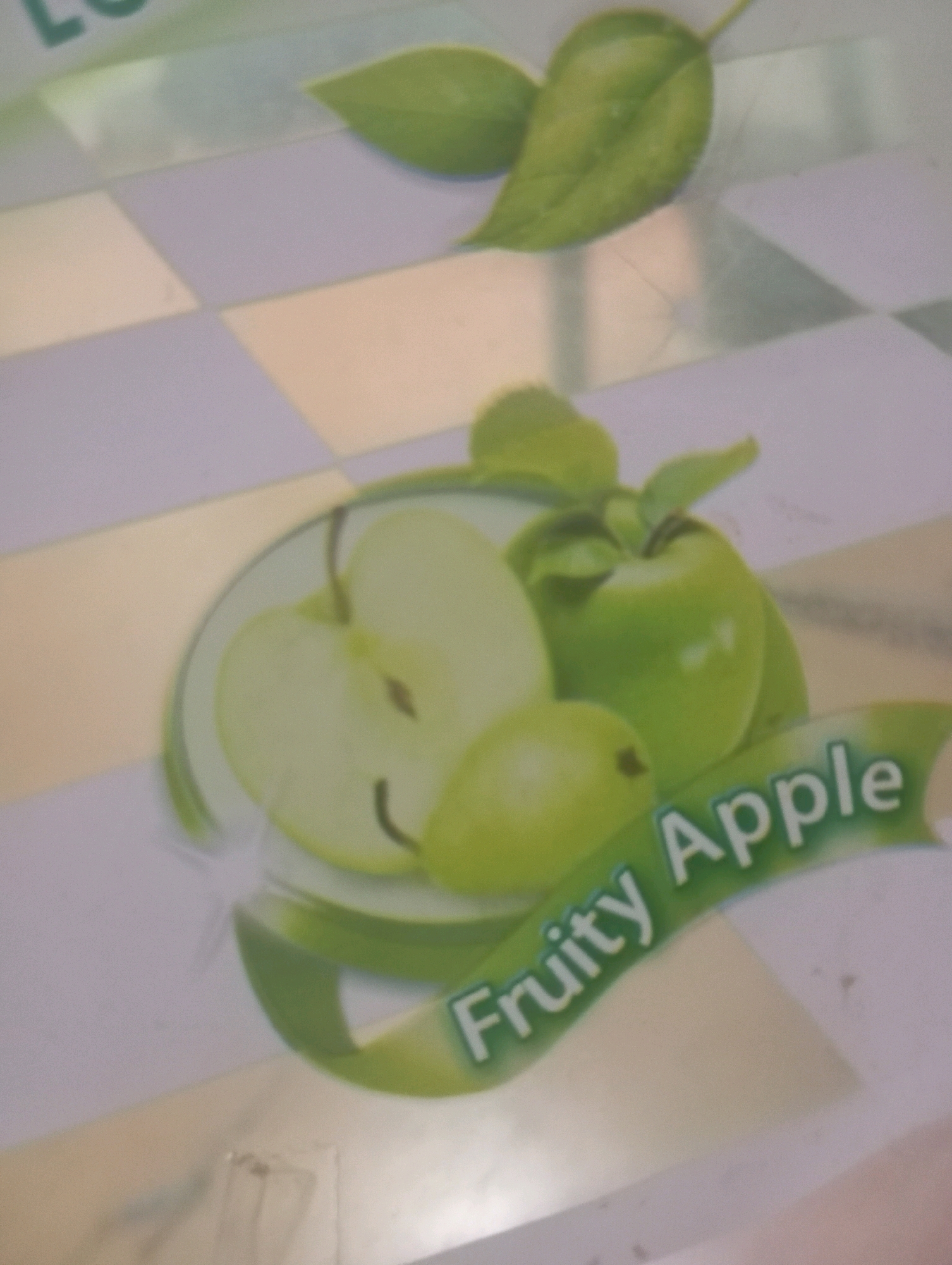 The image shows a close-up of a product label featuring green apples and the text "Fruity Apple". The background is a light-colored checkered pattern. There are no people or discernible environmental details related to Kutasari, Indonesia.