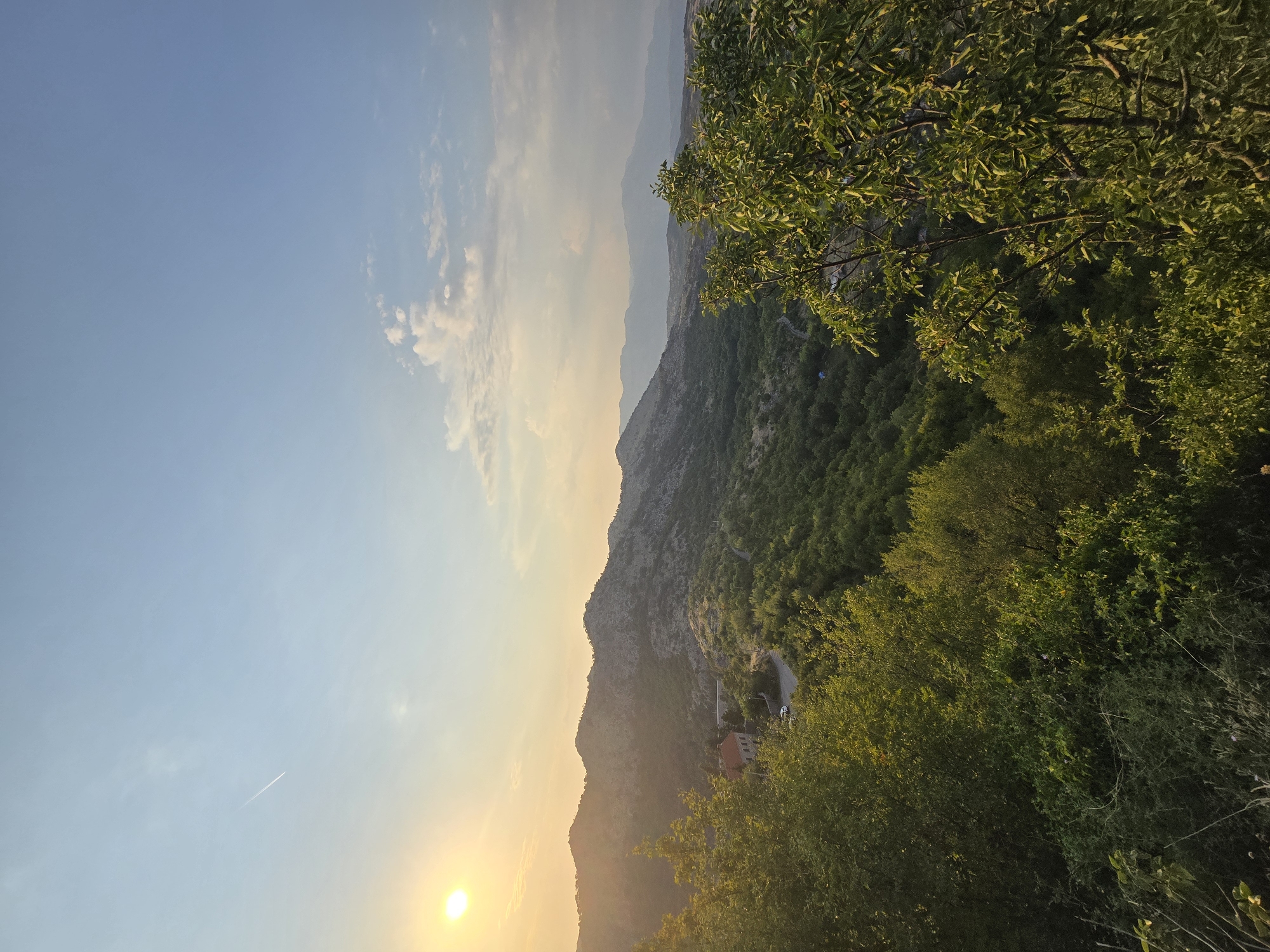 The image captures a scenic view of rolling hills and mountains bathed in the warm glow of the setting sun. The foreground is dominated by lush green trees and vegetation, with a few branches extending into the frame. In the mid-ground, a winding road snakes through the landscape, leading to a small cluster of buildings, one of which appears to be a house with a red roof. The hills are covered in dense green foliage, interspersed with rocky outcrops. The background features a range of mountains fading into the hazy horizon. The sky is a pale blue with scattered white clouds, and the sun, a bright orange orb, is visible in the upper left corner, casting long shadows. A thin contrail from an aircraft is visible in the sky. The overall impression is one of tranquility and natural beauty.