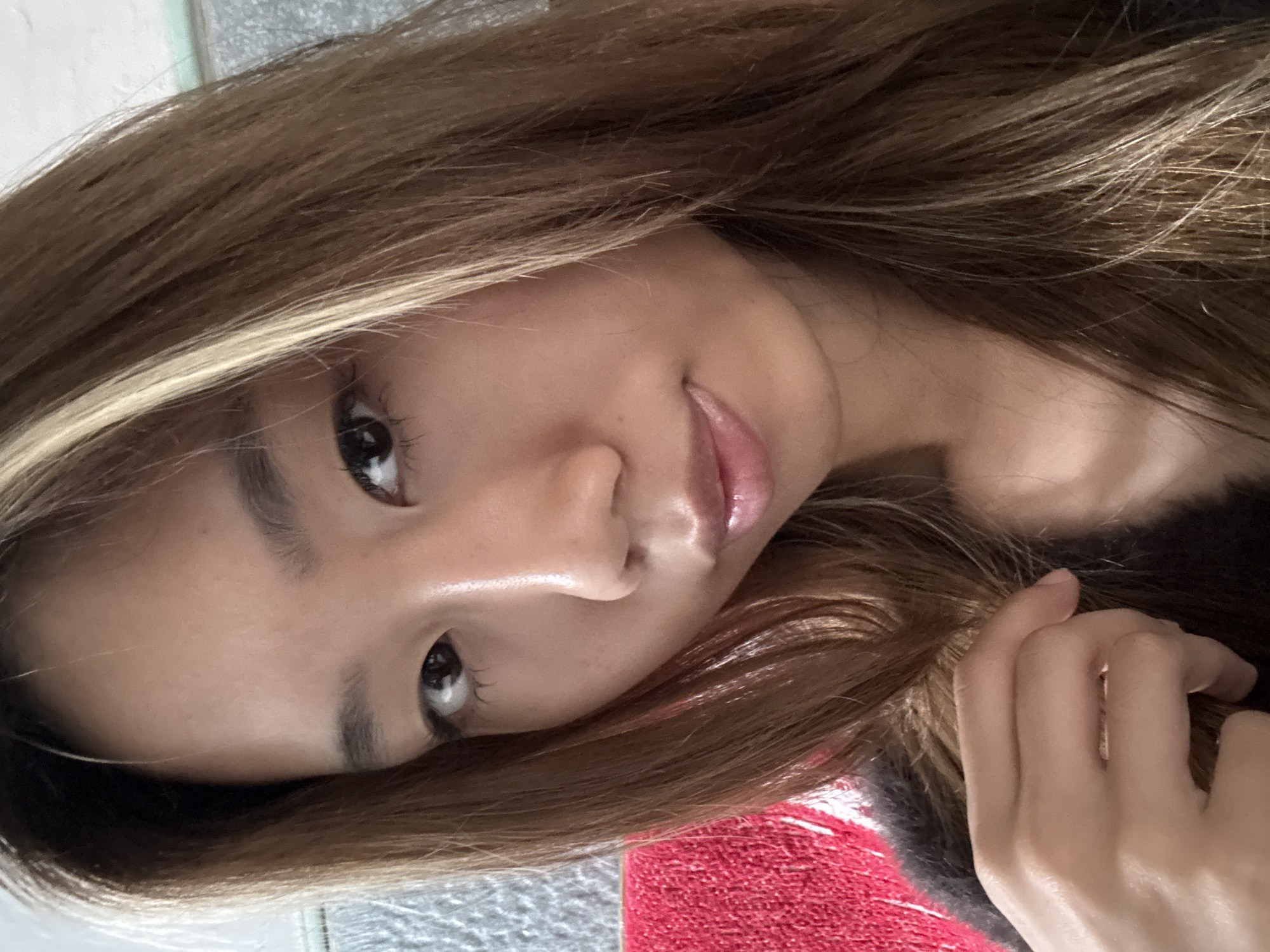 A close-up portrait of a young woman, captured in a selfie-style shot, is presented rotated 90 degrees counter-clockwise. Her head is tilted, and she looks directly into the camera with dark, reflective eyes. Her long, wavy brown hair, featuring prominent lighter blonde or caramel highlights, frames her face and cascades around her shoulders. Her lips are full, glossy, and slightly parted in a gentle, almost enigmatic smile. Her skin appears smooth and well-lit, suggesting indoor lighting. A hand is visible on the lower right side of the frame, subtly touching her hair. She is wearing what appears to be dark, possibly fuzzy, clothing on her right shoulder. The background includes a textured red fabric, possibly a towel or blanket, and a lighter, mottled grey-white surface. The image conveys a calm and intimate mood. There is no visible text.