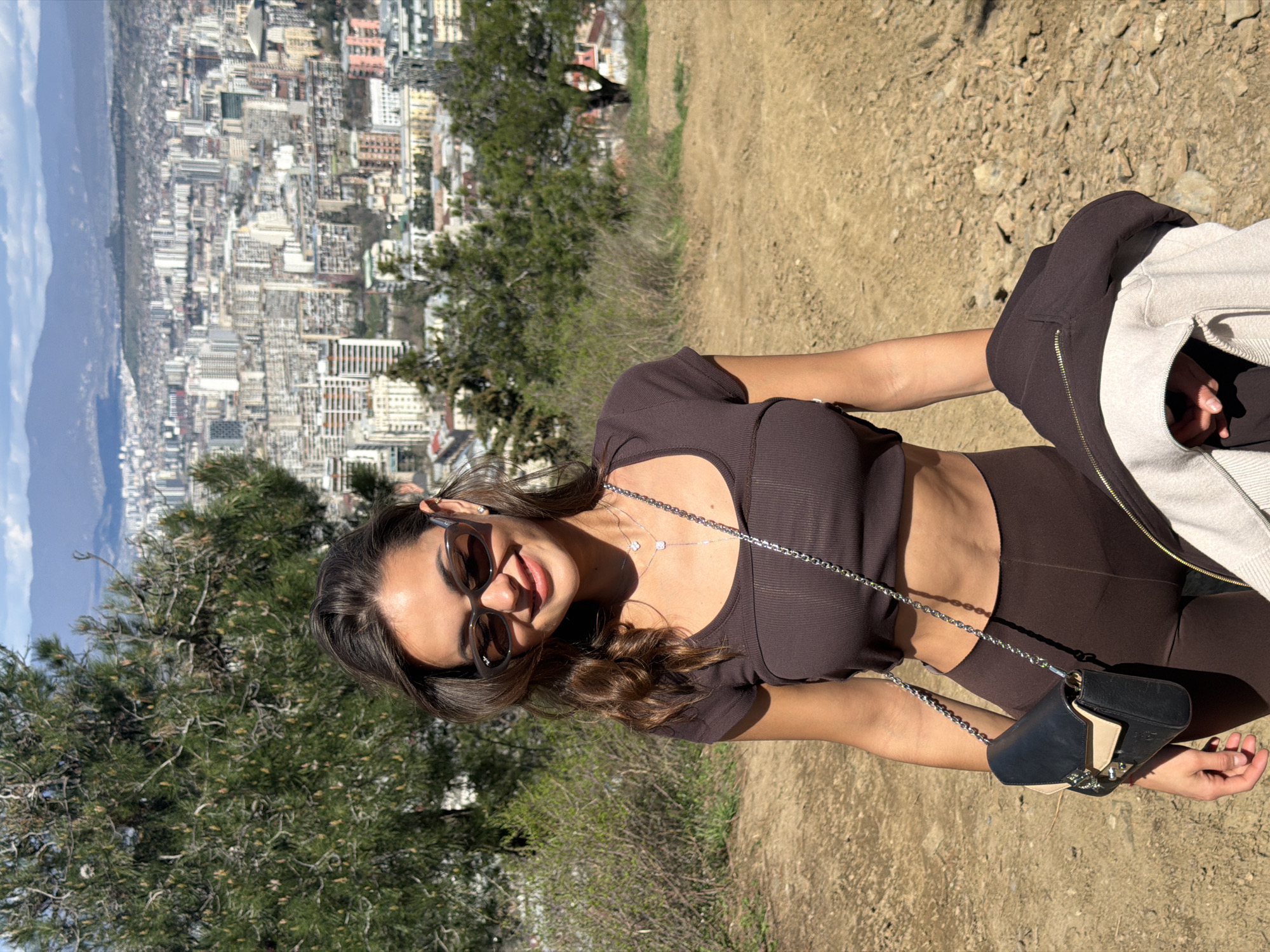 A young woman poses on a path overlooking Tbilisi, Georgia. She wears a brown crop top, leggings, and sunglasses, with a silver chain necklace and a small black purse. A brown and beige jacket is draped over her arm. The background shows a dense cityscape stretching towards mountains under a partly cloudy sky. Vegetation lines the path. The ground is arid and rocky.