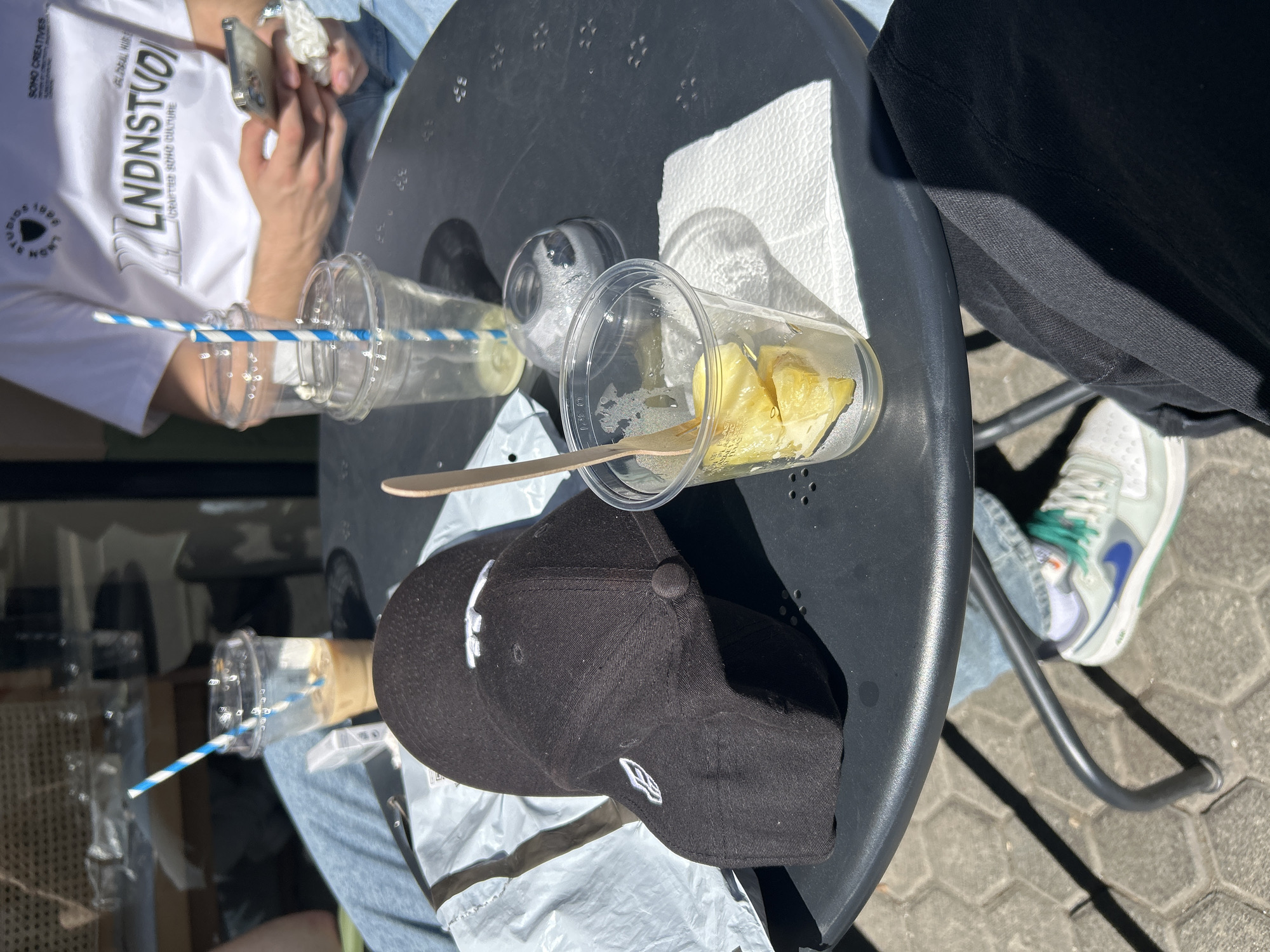 The scene is set on a black metal outdoor table in what appears to be Sofia, Bulgaria based on visual cues. On the table, there are two clear plastic cups with blue and white striped straws, one mostly empty, and the other containing pieces of pineapple with a wooden spoon inside. A black baseball cap with a white logo is placed on a white plastic bag, also on the table. Two people are partially visible: one in a white t-shirt with a black logo holding a cell phone, and another wearing black pants and white and grey sneakers with a blue swoosh. The table is situated on a textured grey surface, likely pavement or patio stones. The lighting suggests a sunny day.